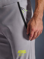 Pantaloni Alpinestars Aeroshell, grigio medio con dettagli giallo fluo, vista ravvicinata della tasca anteriore con zip, bordo nastrato nero e cordini giallo vivace, logo ASTARS giallo sulla coscia, materiale softshell elasticizzato per l'uso in moto.