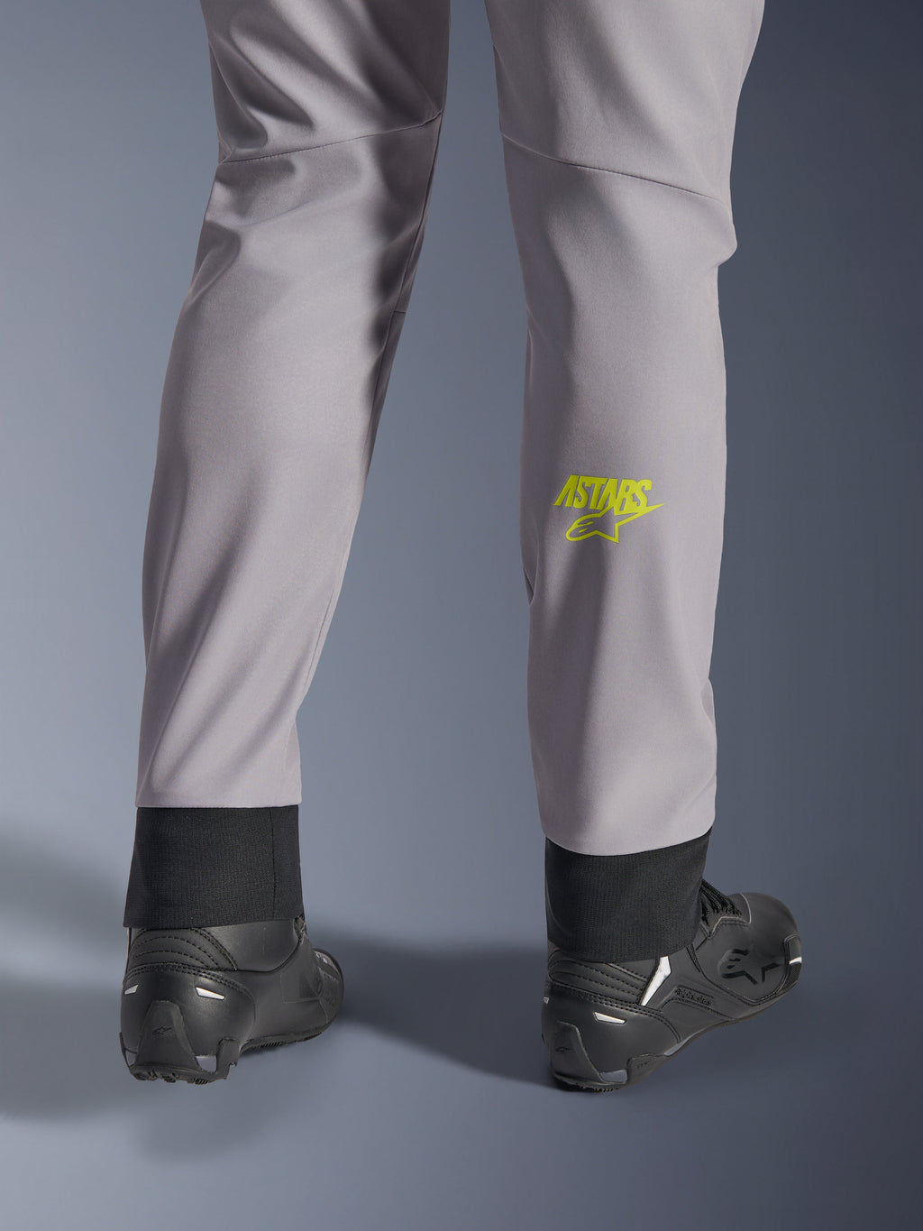 Pantaloni Alpinestars Aeroshell, grigio medio con logo Astars giallo fluo sul polpaccio, vista posteriore con scarpe da moto nere, caratterizzati da orlo inferiore elastico e silhouette moderna affusolata