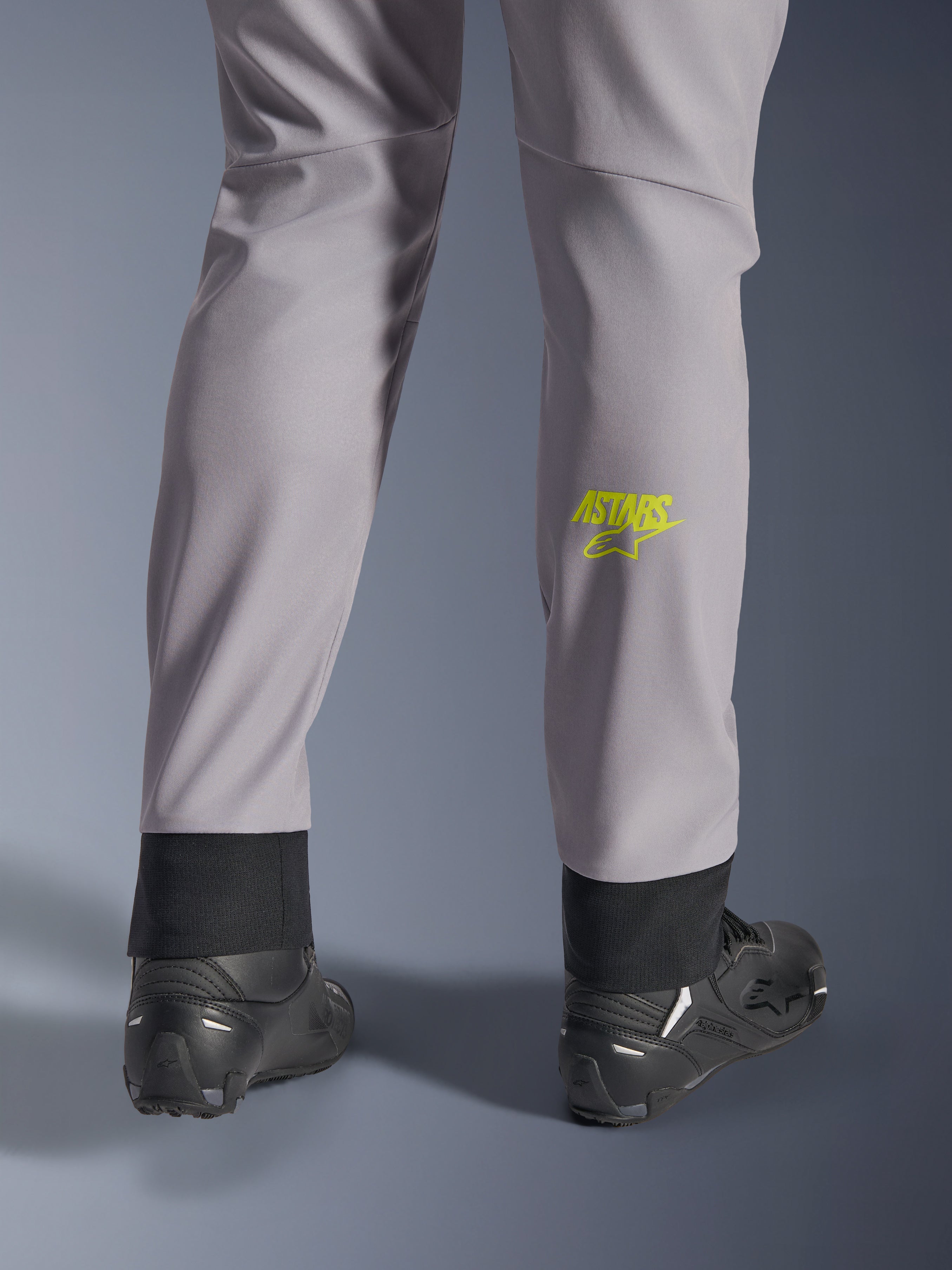 Pantaloni Alpinestars Aeroshell, grigio medio con logo Astars giallo fluo sul polpaccio, vista posteriore con scarpe da moto nere, caratterizzati da orlo inferiore elastico e silhouette moderna affusolata