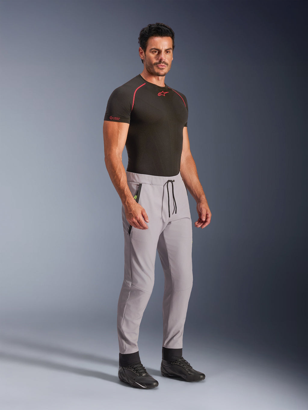 Pantaloni Alpinestars Aeroshell, grigio medio con accenti giallo fluo, pantaloni softshell crossover indossati da un modello con uno strato base nero e scarpe nere, caratterizzati da un taglio moderno affusolato, vita elastica con cordoncino nero e tasche con zip.