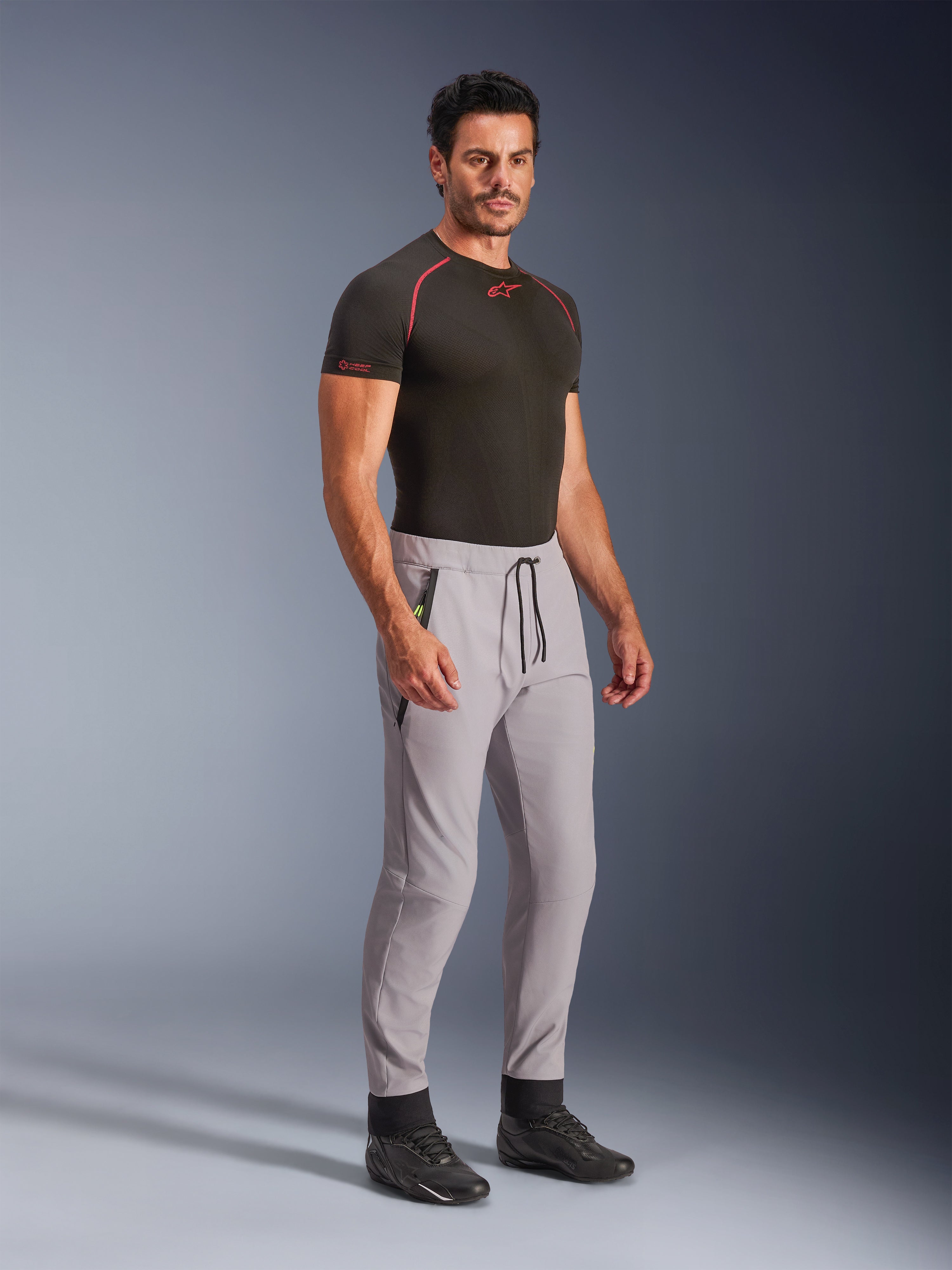Pantaloni Alpinestars Aeroshell, grigio medio con accenti giallo fluo, pantaloni softshell crossover indossati da un modello con uno strato base nero e scarpe nere, caratterizzati da un taglio moderno affusolato, vita elastica con cordoncino nero e tasche con zip.