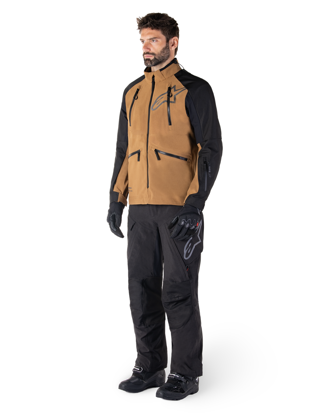 Alpinestars Hyde XT Drystar® Pants, pantaloni da moto adventure, nero, indossati con giacca tecnica marrone e nera, guanti e stivali neri, caratterizzati da un design sopra lo stivale per la guida off-road e dual-sport
