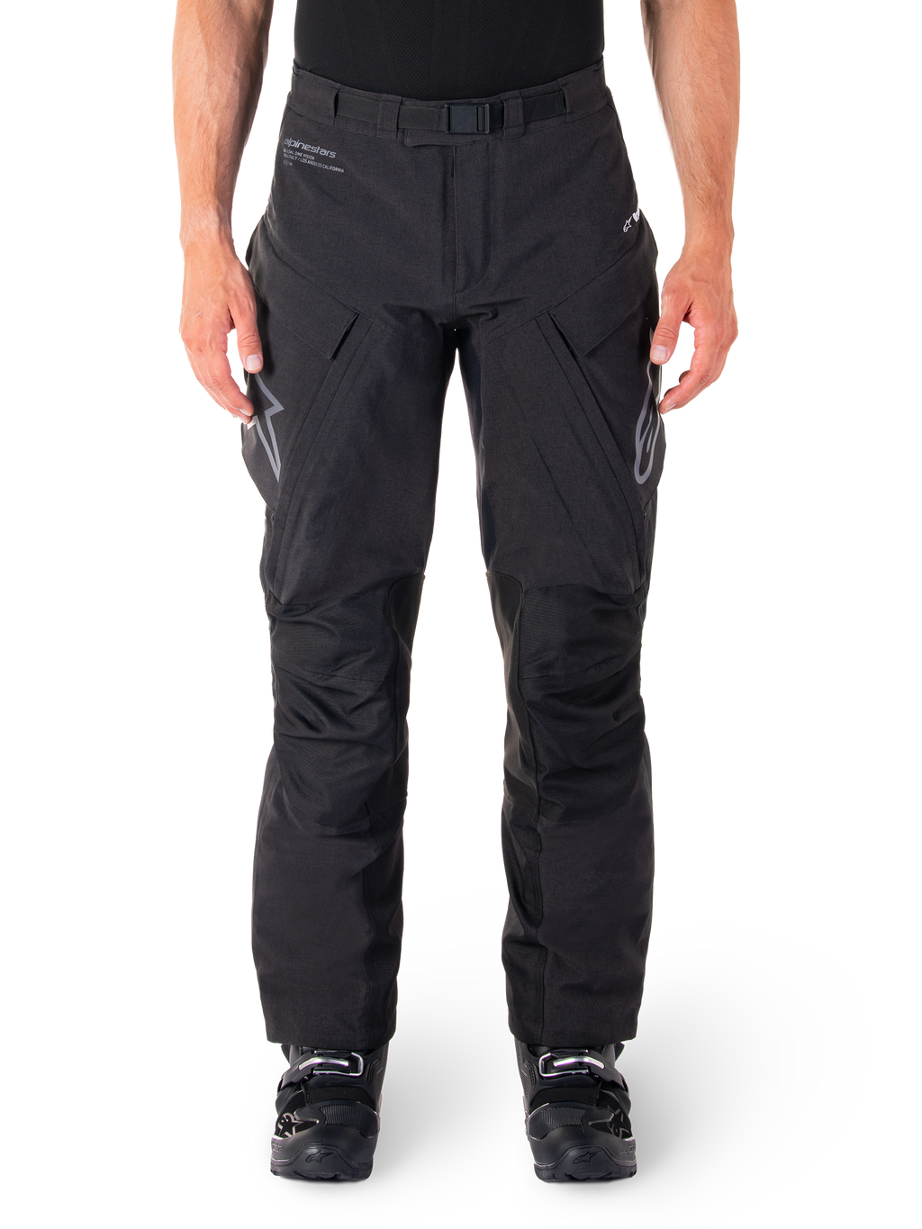Pantaloni Alpinestars Hyde XT Drystar, pantaloni adventure touring neri, indossati da un pilota con stivali neri, con loghi grigi sulle cosce, cintura integrata, pannelli rinforzati sulle ginocchia e costruzione in tessuto impermeabile