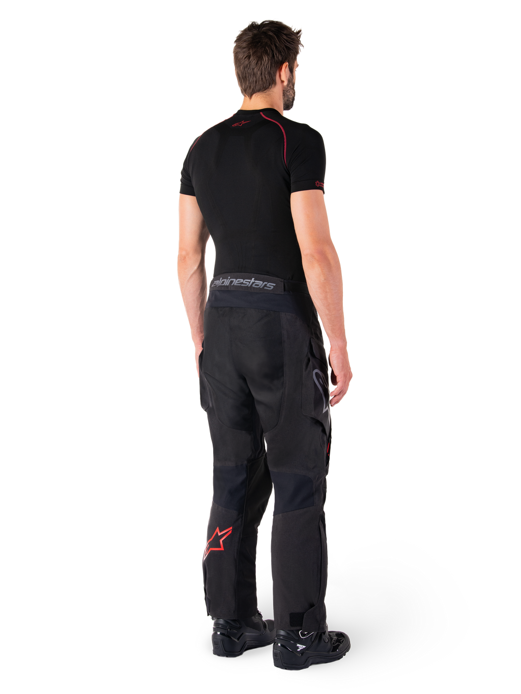 Pantaloni Alpinestars Hyde XT Drystar®, pantaloni Adventure Touring, nero e rosso vivace, vista posteriore di un pilota che indossa i pantaloni con uno strato di base nero e stivali neri, caratterizzati da una robusta struttura in tessuto con logo a stella rosso sulla parte inferiore della gamba e marchio Alpinestars grigio sulla fascia in vita
