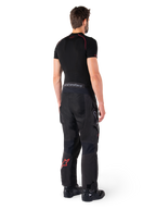 Pantaloni Alpinestars Hyde XT Drystar®, pantaloni Adventure Touring, nero e rosso vivace, vista posteriore di un pilota che indossa i pantaloni con uno strato di base nero e stivali neri, caratterizzati da una robusta struttura in tessuto con logo a stella rosso sulla parte inferiore della gamba e marchio Alpinestars grigio sulla fascia in vita