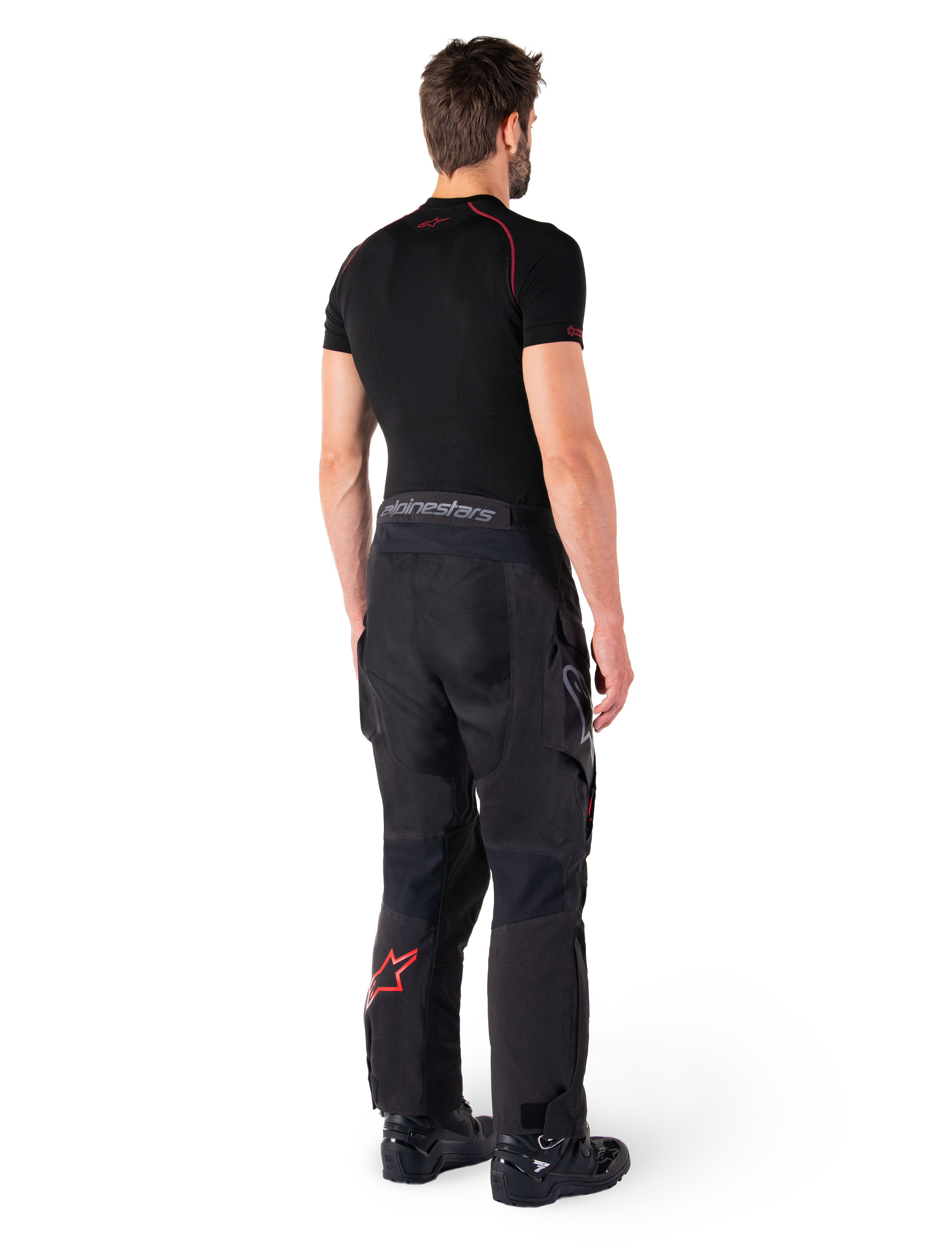 Pantaloni Alpinestars Hyde XT Drystar®, pantaloni Adventure Touring, nero e rosso vivace, vista posteriore di un pilota che indossa i pantaloni con uno strato di base nero e stivali neri, caratterizzati da una robusta struttura in tessuto con logo a stella rosso sulla parte inferiore della gamba e marchio Alpinestars grigio sulla fascia in vita