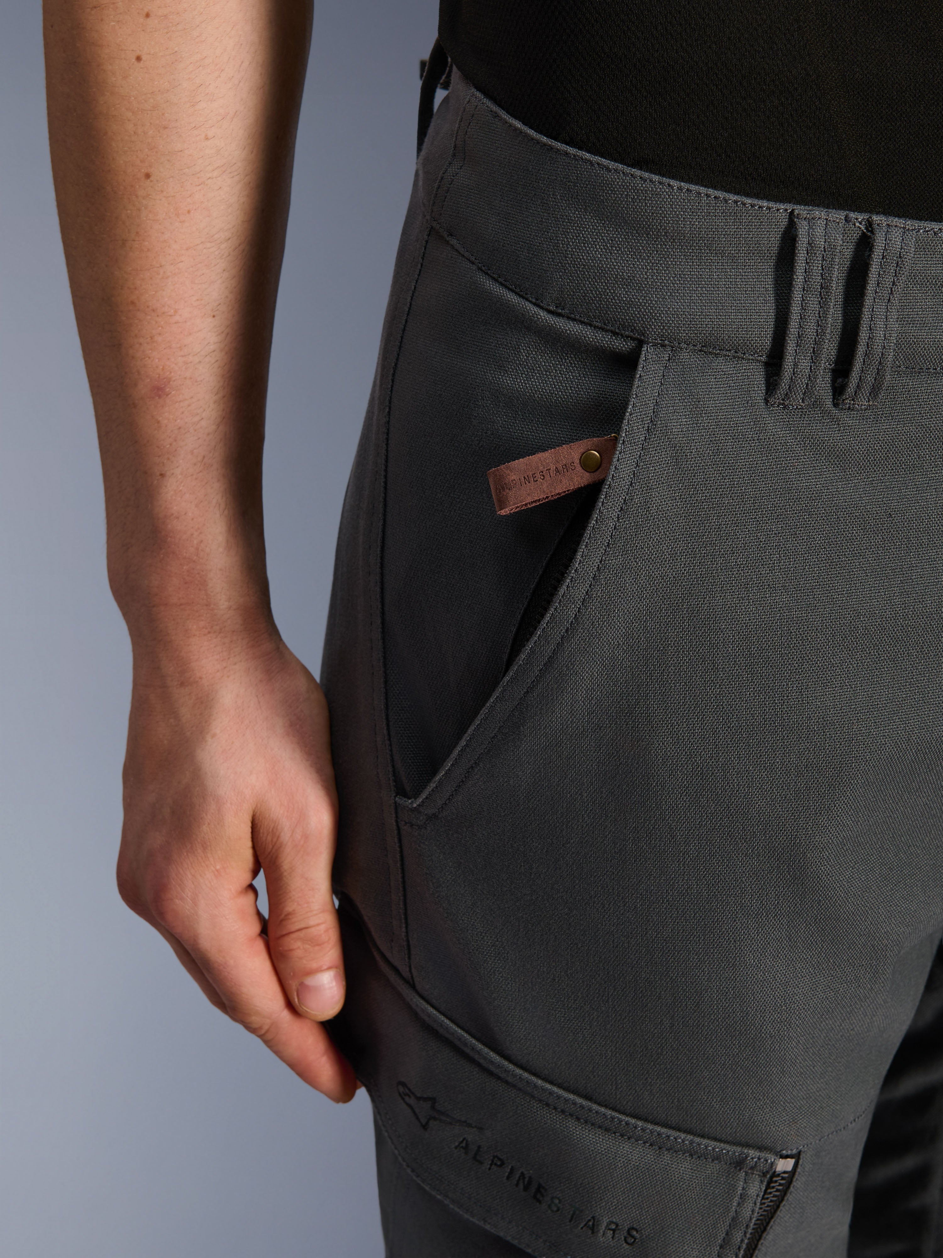 Alpinestars Borrego Drystar® Pants, pantaloni da moto adventure, grigio antracite, dettaglio ravvicinato della tasca e del fianco, texture in tela cerata con etichetta in pelle marrone con logo e marchio Alpinestars ricamato sulla tasca cargo della coscia.