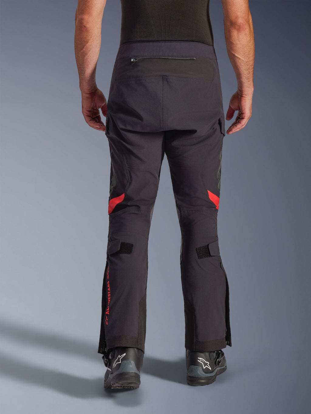 Alpinestars Halo Pro Drystar® XF Laminated Pants, Black Dark Shadow, vista posteriore di pantaloni da moto adventure con accenti rossi dietro le ginocchia e logo rosso sul polpaccio, indossati con stivali Alpinestars neri, tessuto laminato 3L resistente per prestazioni su ogni terreno