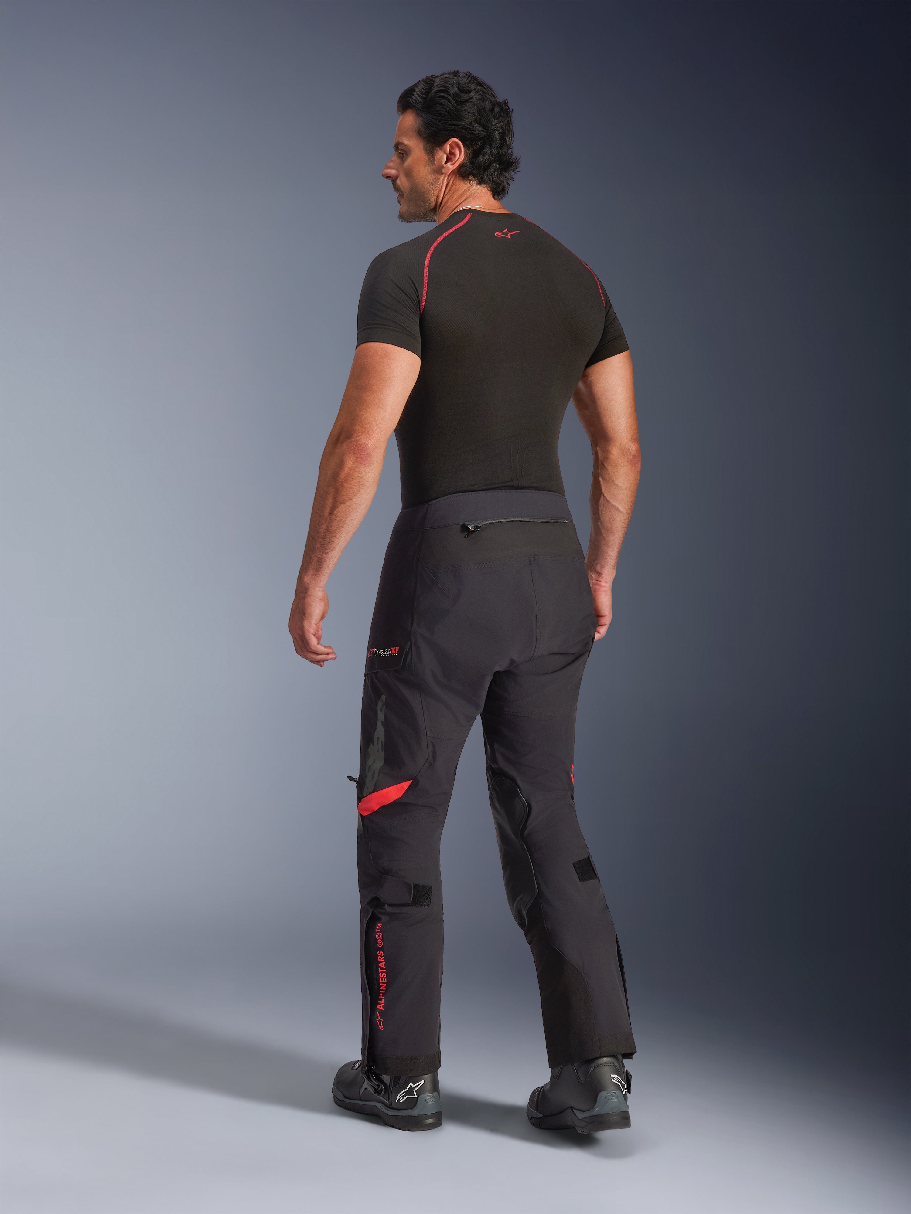Alpinestars Halo Pro Drystar® XF Pantaloni Laminati, Nero Dark Shadow con accenti rossi, vista posteriore su modello, abbinati a base layer nero e stivali da moto, abbigliamento tecnico da adventure touring per ogni terreno