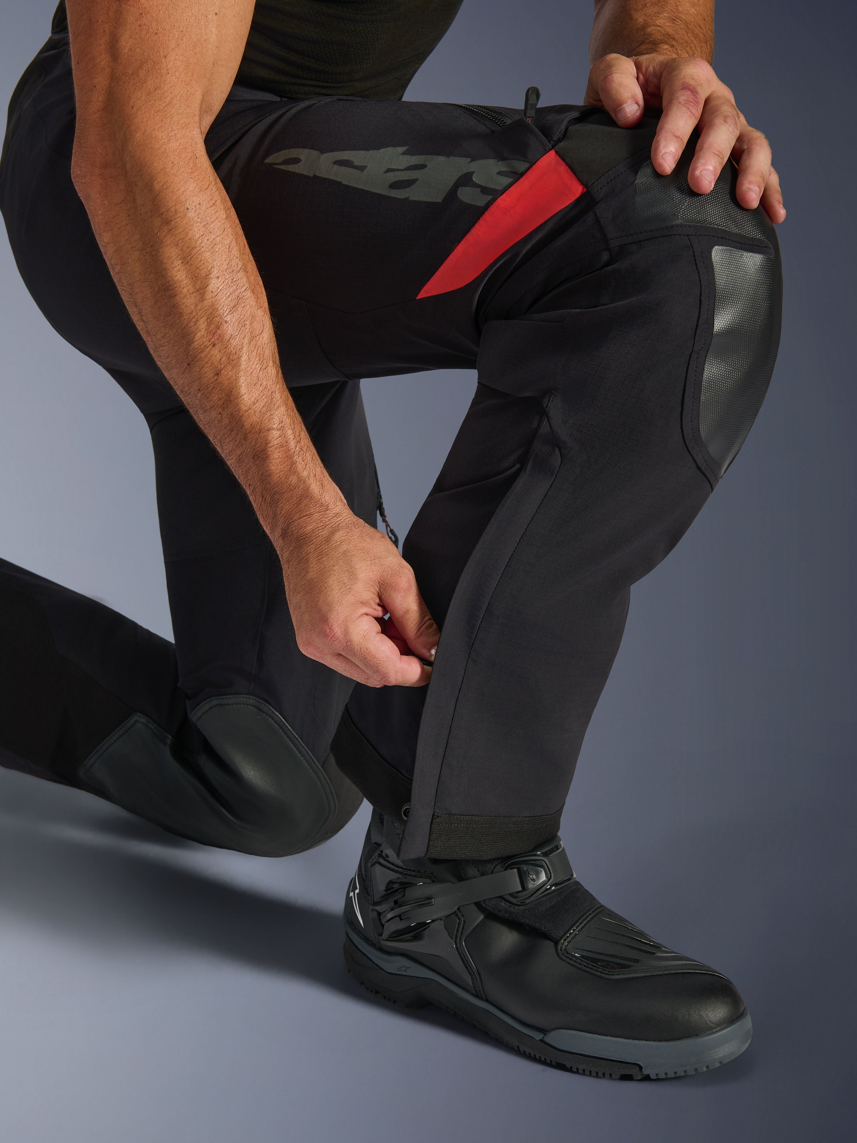 Alpinestars Halo Pro Drystar® XF Laminated Pants, Black Dark Shadow, primo piano della parte inferiore della gamba con cerniera regolabile sul polpaccio, caratterizzati da rinforzi durevoli sulle ginocchia, dettagli rossi e logo 'astars', indossati con stivali da moto neri.