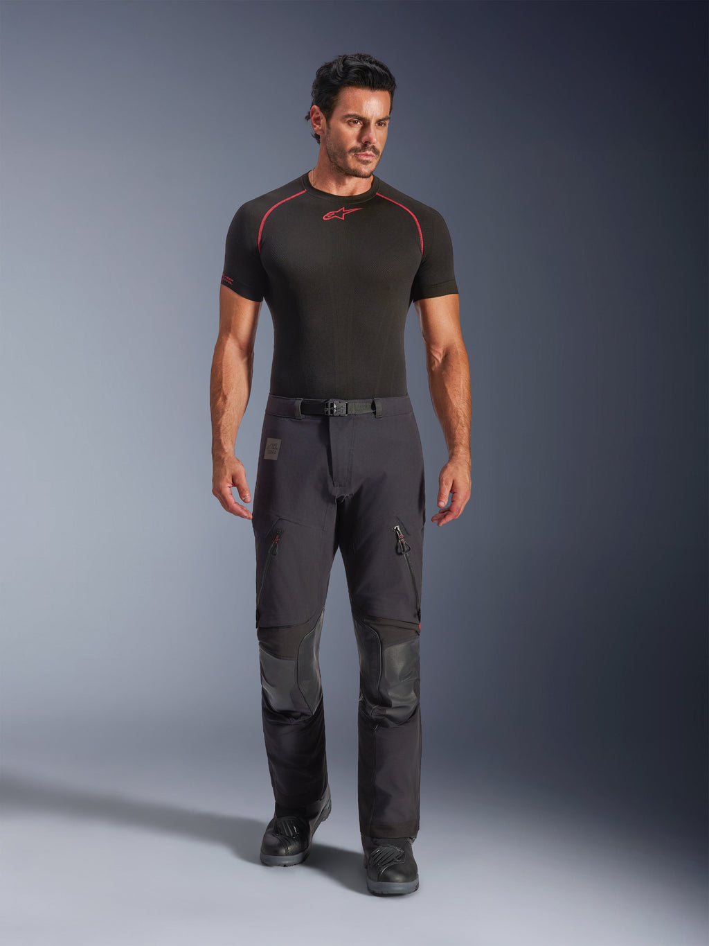 Pantaloni laminati Alpinestars Halo Pro Drystar® XF in Black Dark Shadow, indossati da un modello con uno strato di base nero e stivali da moto, con prese d'aria tecniche sulle cosce con zip, tasche cargo e pannelli rinforzati sulle ginocchia, abbigliamento da moto adventure touring.