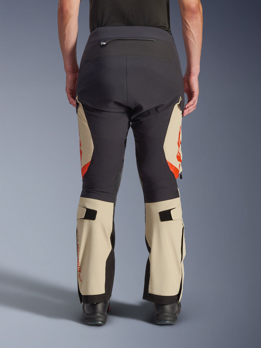 Vista posteriore dei pantaloni laminati Alpinestars Halo Pro Drystar® XF, Mastic Black con pannelli tan e accenti arancioni, pantaloni da moto adventure con costruzione in tessuto tecnico, indossati con stivali da moto neri