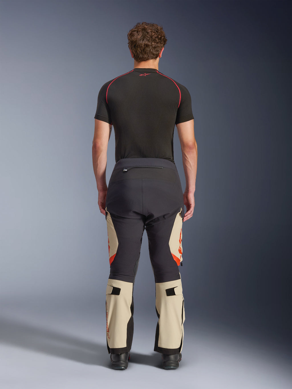Pantaloni laminati Alpinestars Halo Pro Drystar® XF, Mastic Black, vista posteriore su un modello, design a blocchi di colore nero e beige con loghi arancioni, pantaloni da moto adventure touring con robusta struttura in tessuto e sezioni regolabili sui polpacci