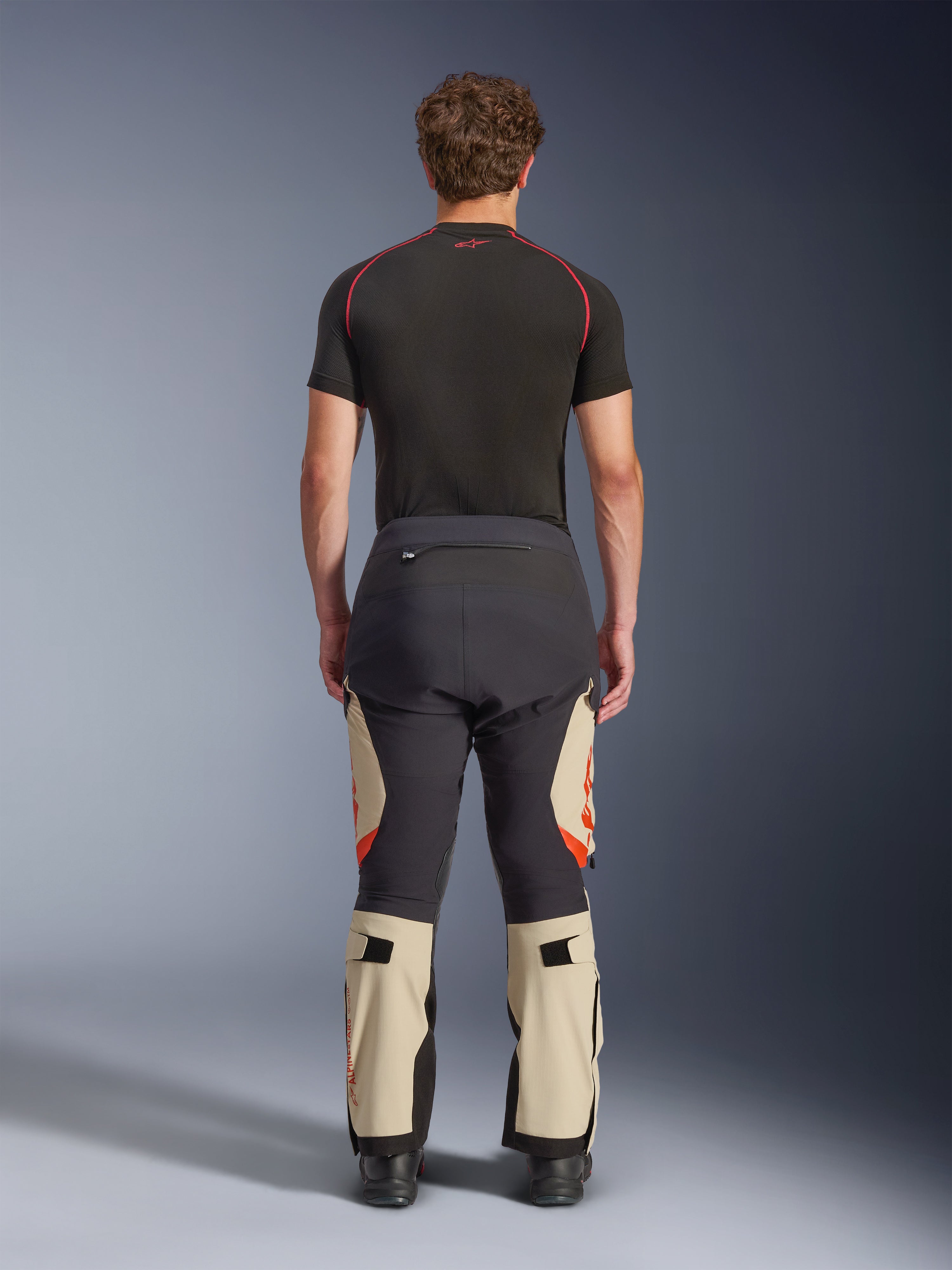 Pantaloni laminati Alpinestars Halo Pro Drystar® XF, Mastic Black, vista posteriore su un modello, design a blocchi di colore nero e beige con loghi arancioni, pantaloni da moto adventure touring con robusta struttura in tessuto e sezioni regolabili sui polpacci