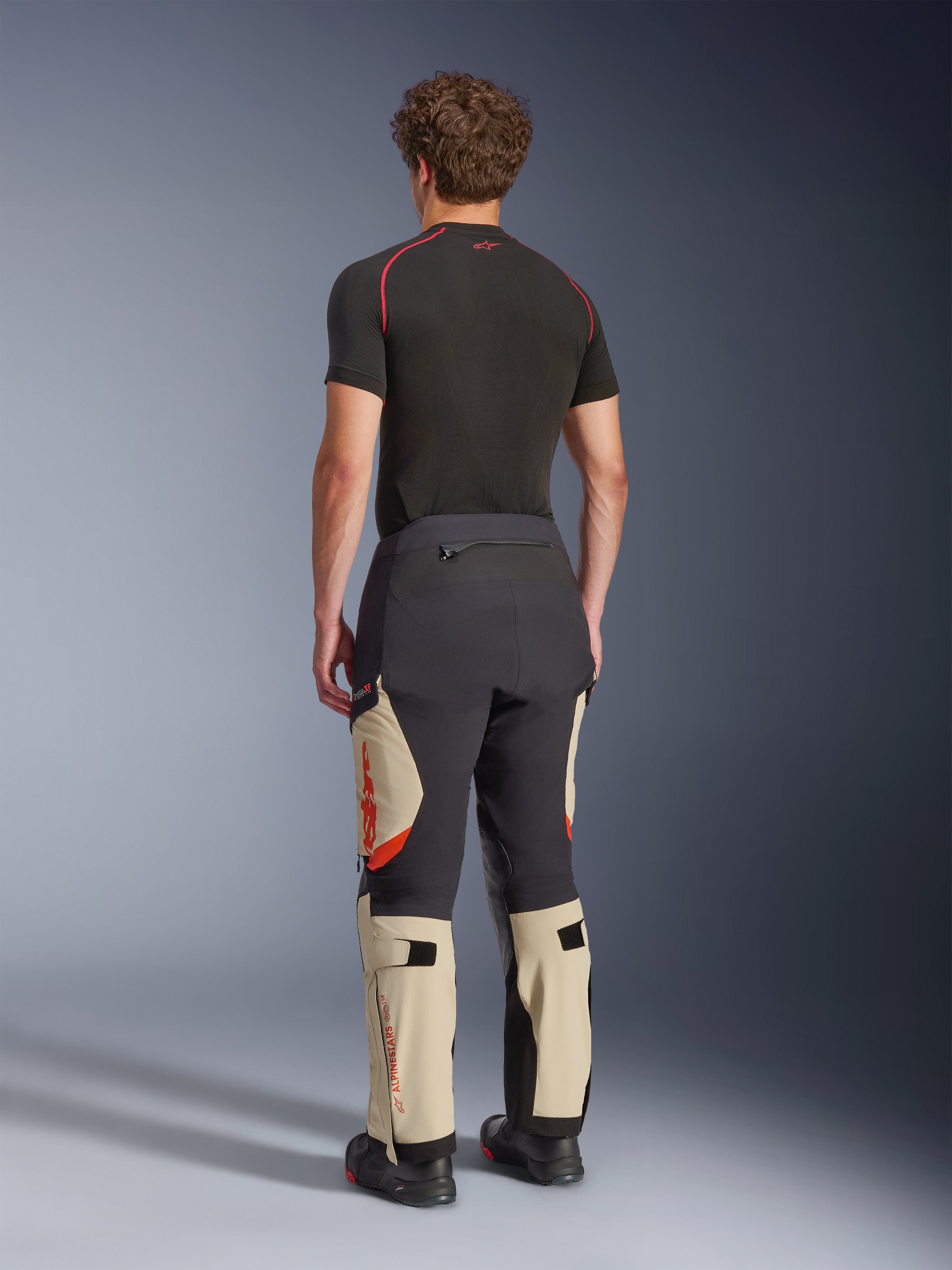 Pantaloni laminati Alpinestars Halo Pro Drystar® XF, pantaloni da moto adventure, Mastic Black e Tan con accenti arancioni, vista posteriore che mostra la costruzione del guscio laminato a 3 strati, abbinati a un base layer nero e stivali adventure.