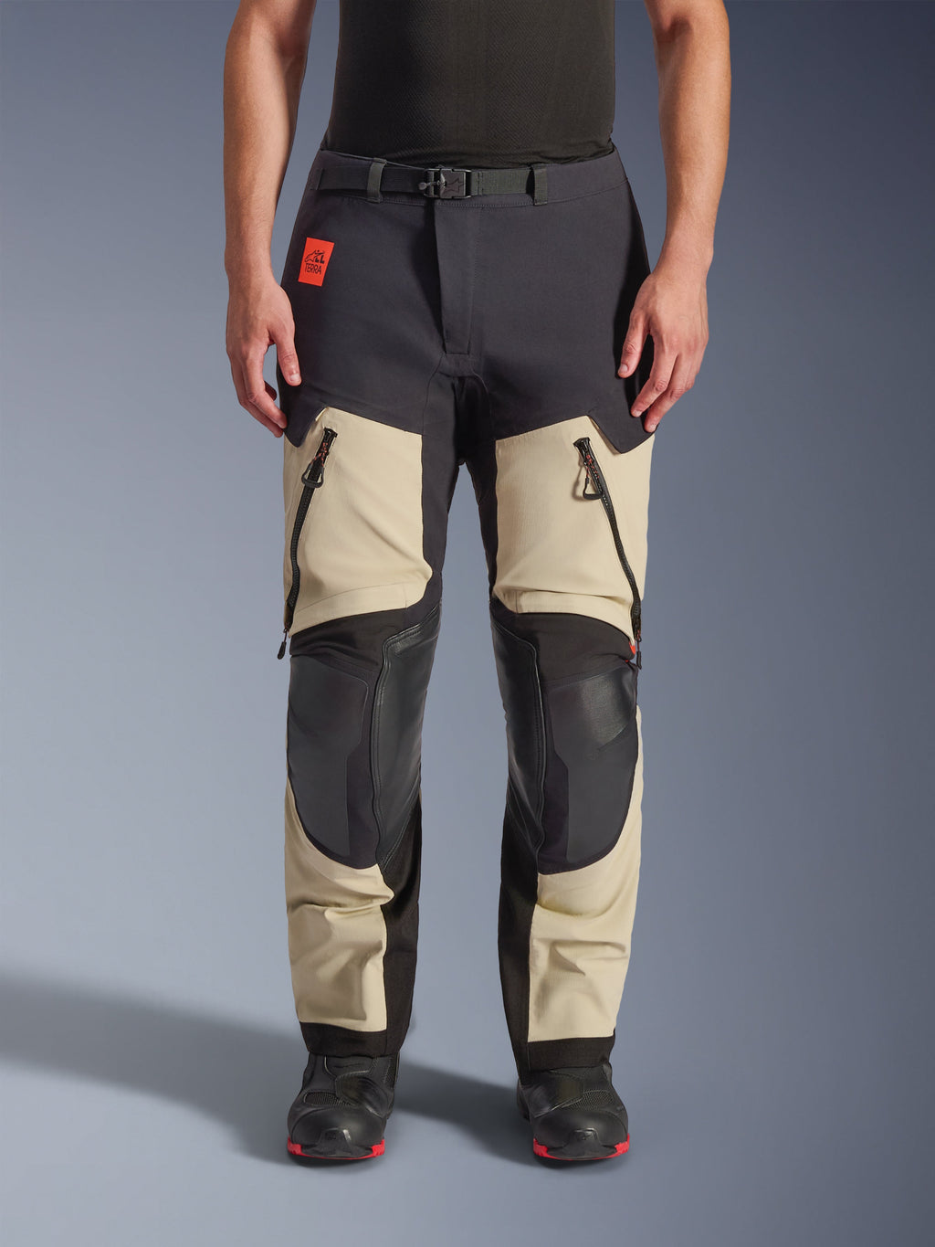 Alpinestars Halo Pro Drystar® XF Laminated Pants, pantaloni da moto adventure, Mastic Black, vista frontale indossati da un pilota, con ampie prese d'aria con cerniera sulle cosce, pannelli rinforzati neri sulle ginocchia e costruzione in tessuto color sabbia, abbinati a stivali da moto neri e rossi