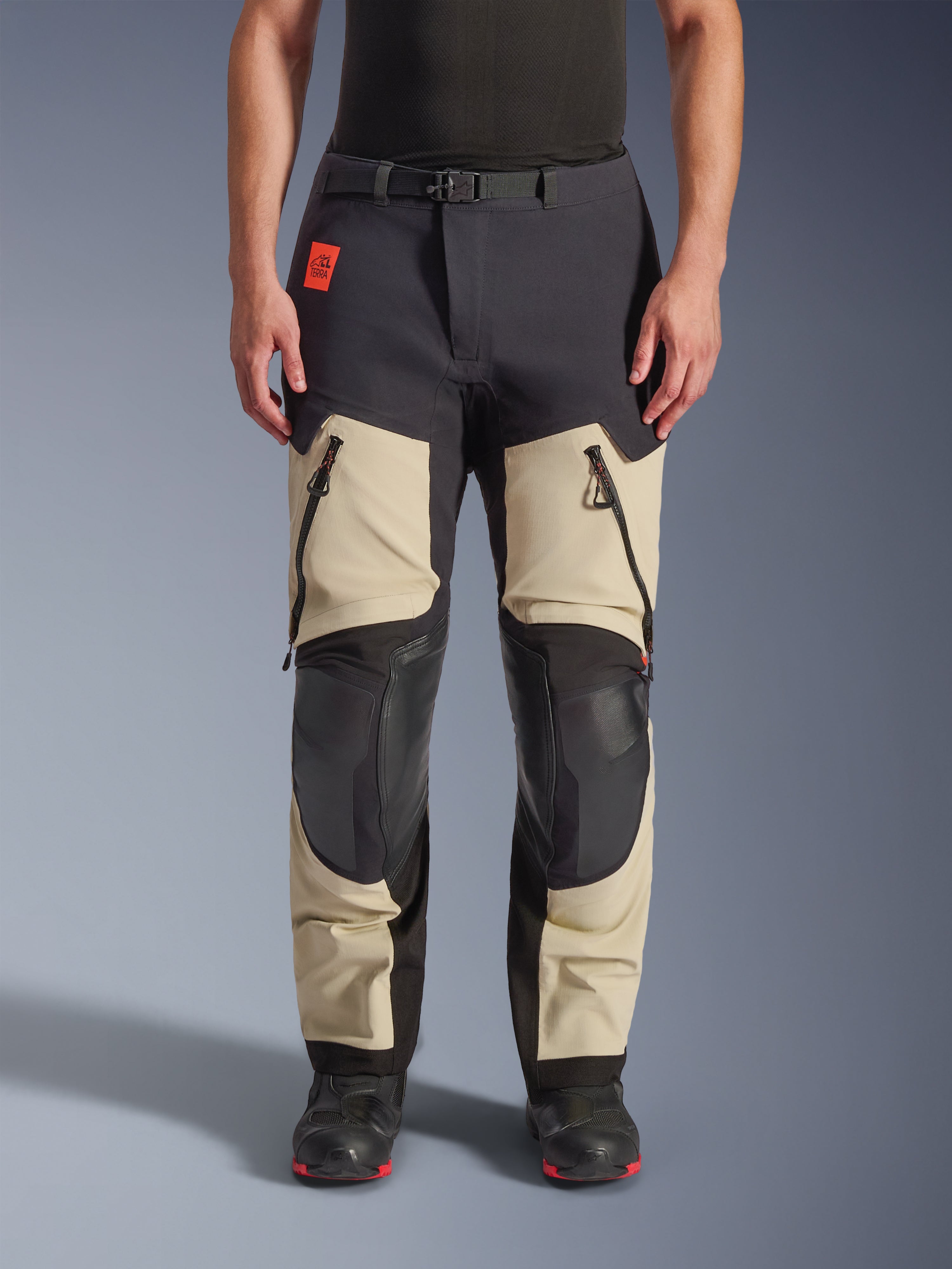 Alpinestars Halo Pro Drystar® XF Laminated Pants, pantaloni da moto adventure, Mastic Black, vista frontale indossati da un pilota, con ampie prese d'aria con cerniera sulle cosce, pannelli rinforzati neri sulle ginocchia e costruzione in tessuto color sabbia, abbinati a stivali da moto neri e rossi