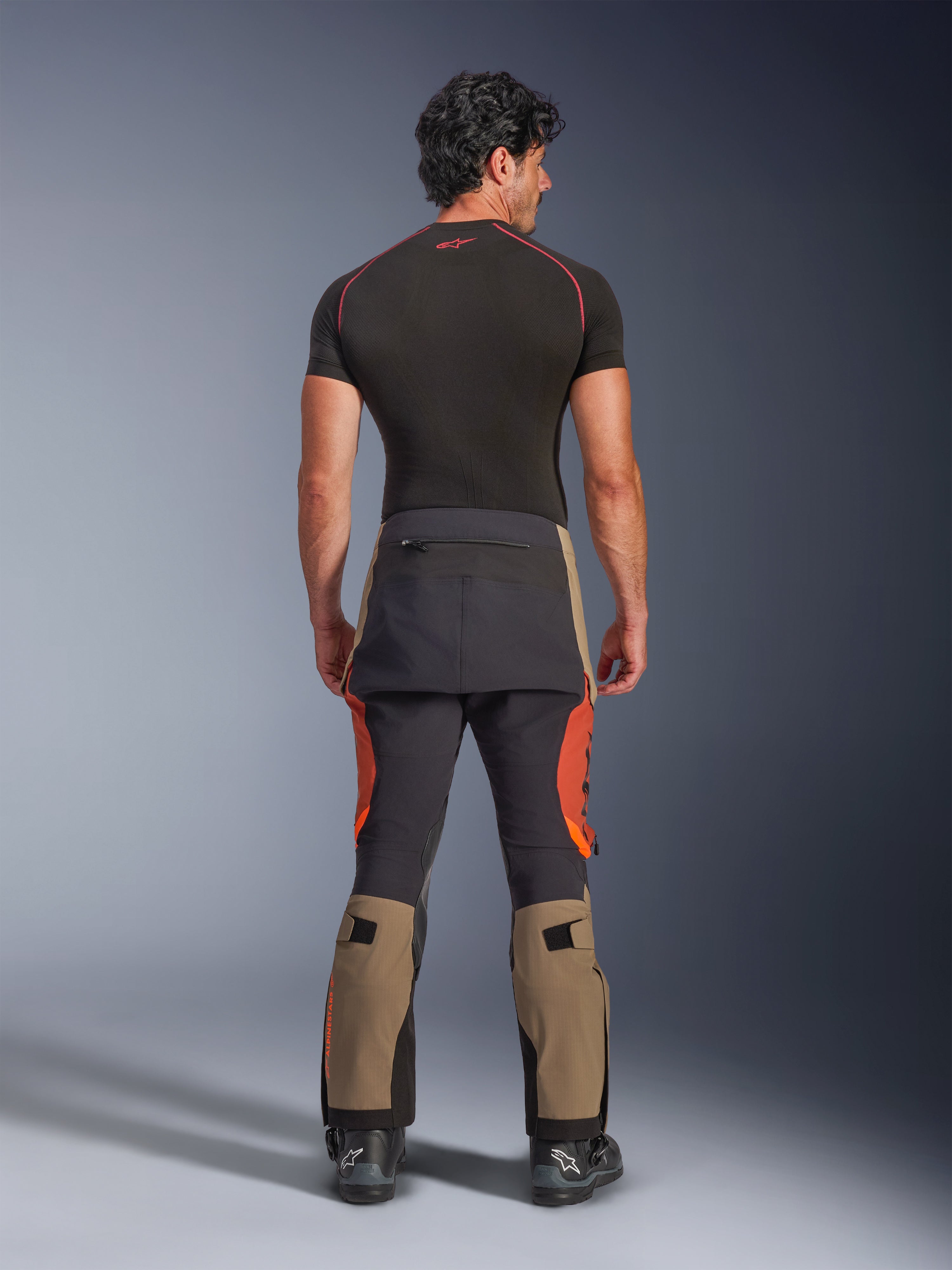 Alpinestars Halo Pro Drystar® XF Laminated Pants, pantaloni da moto adventure Walnut Rust Black, vista posteriore su un modello che indossa stivali neri e un base layer a maniche corte, con parte inferiore delle gambe color noce, accenti arancione ruggine e pannelli rinforzati neri.