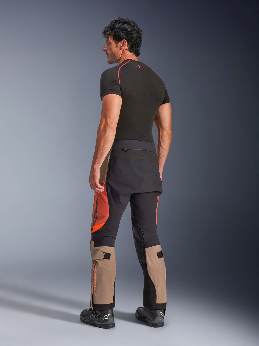 Pantaloni laminati Alpinestars Halo Pro Drystar® XF in Walnut Rust Black, vista posteriore di un modello maschile che indossa pantaloni da moto adventure con pannelli color sabbia, arancione e nero, abbinati a un base layer nero a maniche corte e stivali da moto neri, mettendo in evidenza l'abbigliamento tecnico da guida outdoor