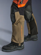 Pantaloni laminati Alpinestars Halo Pro Drystar® XF, pantaloni da moto adventure, Walnut Rust e nero, dettaglio dei pannelli rinforzati sulle ginocchia e del logo arancione, indossati con stivali da touring neri, costruzione in tessuto resistente per guida su ogni terreno