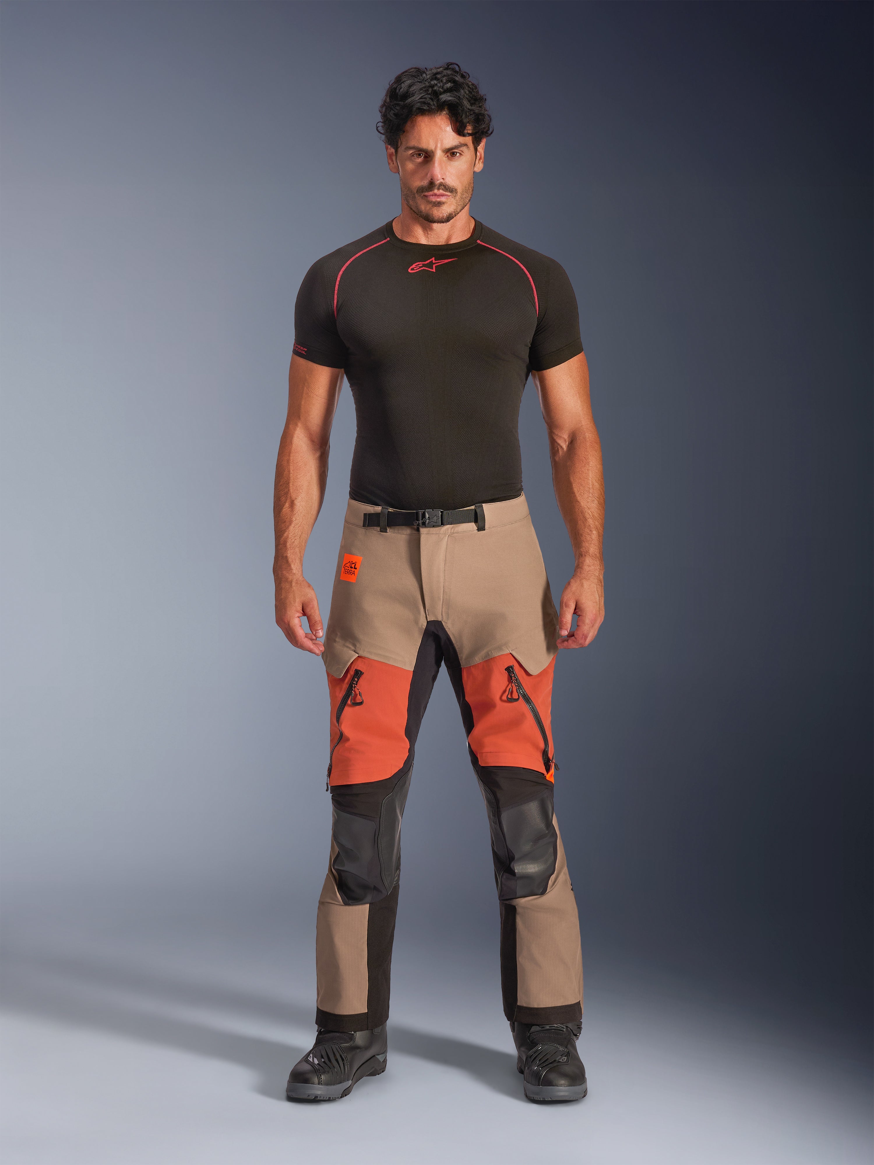 Modello maschile che indossa i pantaloni laminati Alpinestars Halo Pro Drystar® XF, pantaloni da moto Adventure, Walnut Rust Black, design a blocchi di colore beige e arancione con pannelli tecnici neri, caratterizzati da prese d'aria sulle cosce con zip e fibbia magnetica in vita, abbinati a un base layer nero e stivali da moto neri.