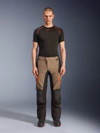 Modello che indossa i pantaloni Alpinestars Andes V4 Drystar® in Walnut Black, pantaloni da moto adventure abbinati a un base layer nero e stivali, con dettagli di cerniere arancioni sulle cosce e pannelli neri rinforzati per una resistenza su tutti i terreni.