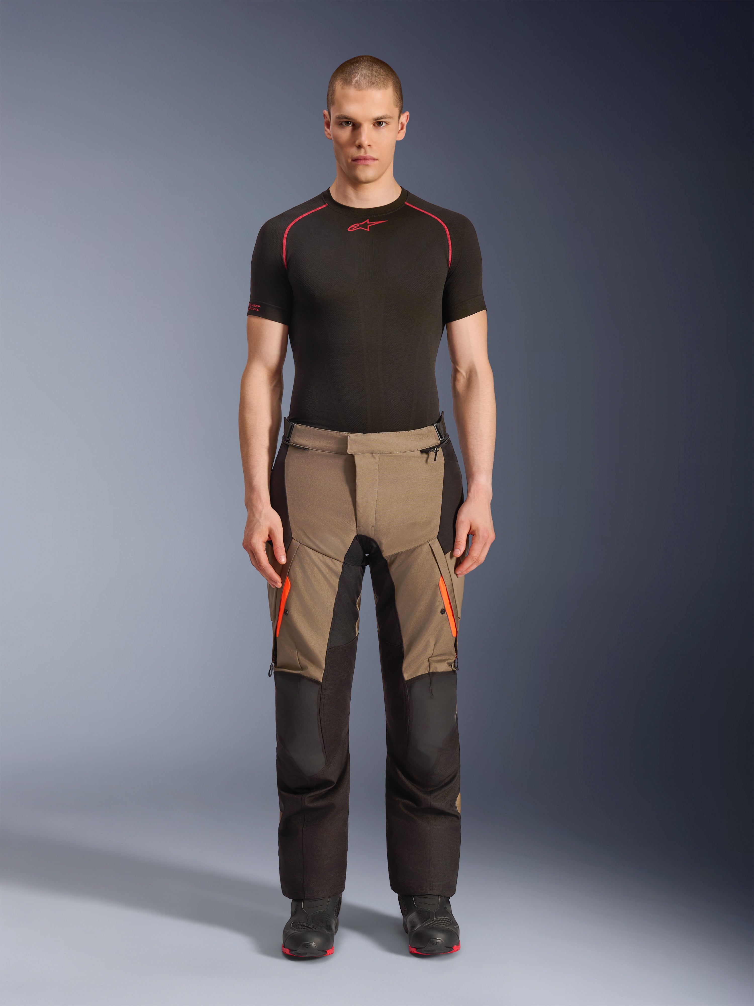 Modello che indossa i pantaloni Alpinestars Andes V4 Drystar® in Walnut Black, pantaloni da moto adventure abbinati a un base layer nero e stivali, con dettagli di cerniere arancioni sulle cosce e pannelli neri rinforzati per una resistenza su tutti i terreni.