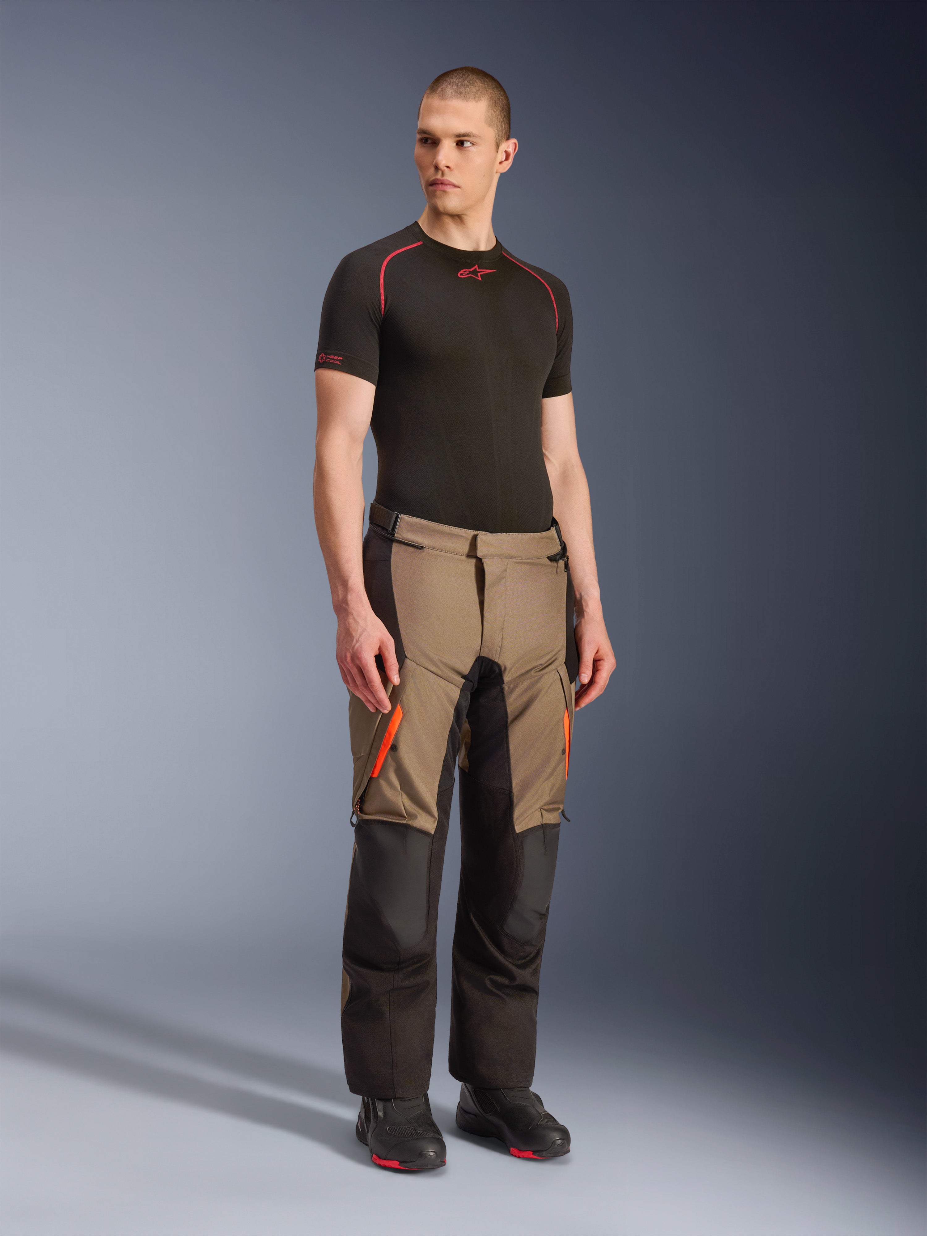 Alpinestars Andes V4 Drystar® Pants, pantaloni da moto adventure touring, Walnut Black con accenti arancioni, indossati da un modello con base layer nero a maniche corte e stivali neri, con pannelli in tessuto rinforzato e tasche cargo, design robusto per la guida in ogni condizione atmosferica