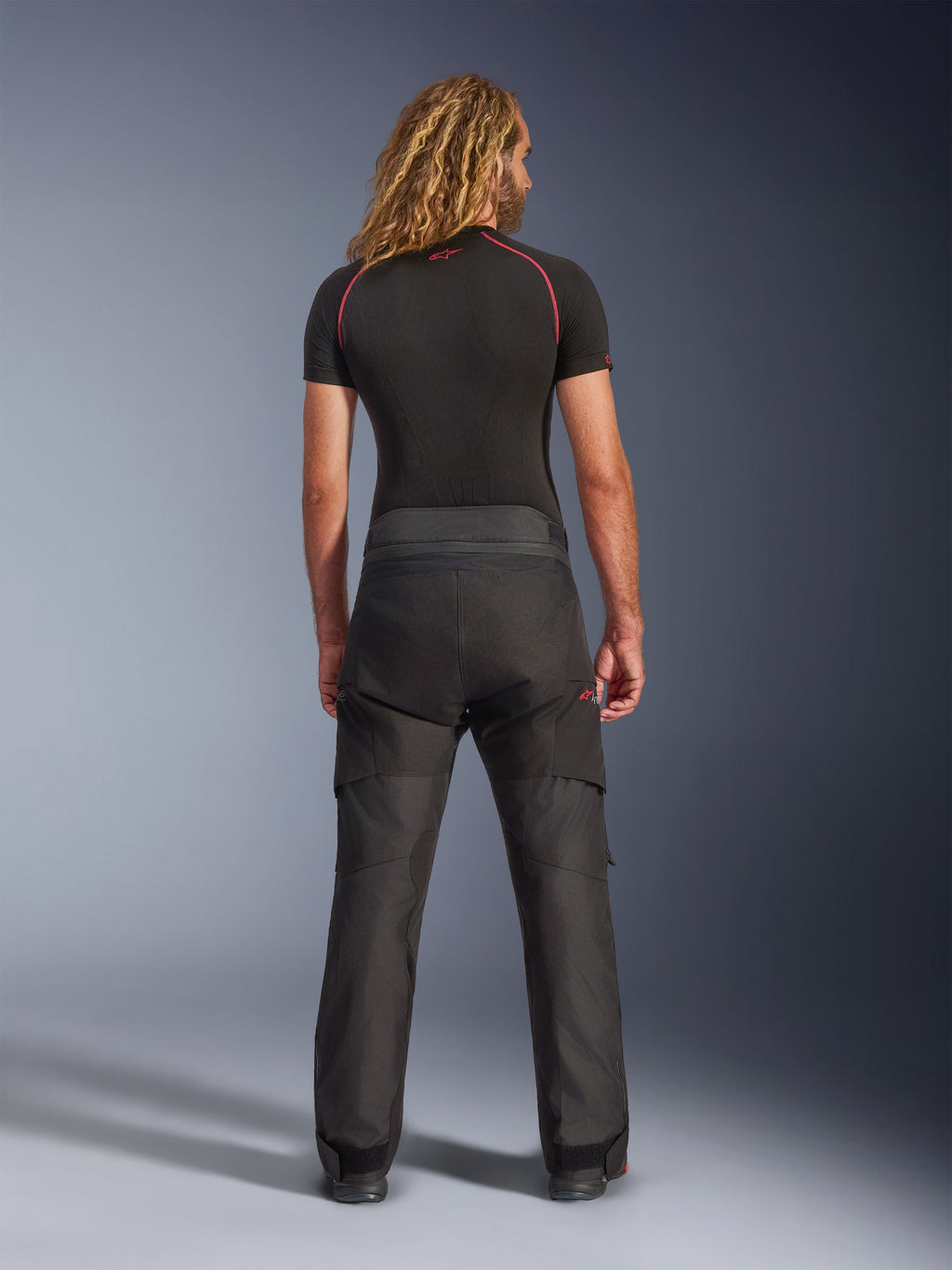Vista posteriore di un modello che indossa i pantaloni Alpinestars Andes V4 Drystar® - Short, pantaloni da moto adventure neri, abbinati a un base layer nero con cuciture rosse, che mostrano la costruzione in tessuto resistente e la vestibilità ergonomica.