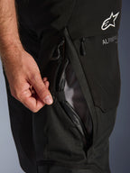 Pantaloni Alpinestars Andes V4 Drystar® neri, primo piano di una mano che apre la presa d'aria diretta sulla coscia con fodera in rete, dettaglio della robusta costruzione in tessuto e branding Alpinestars