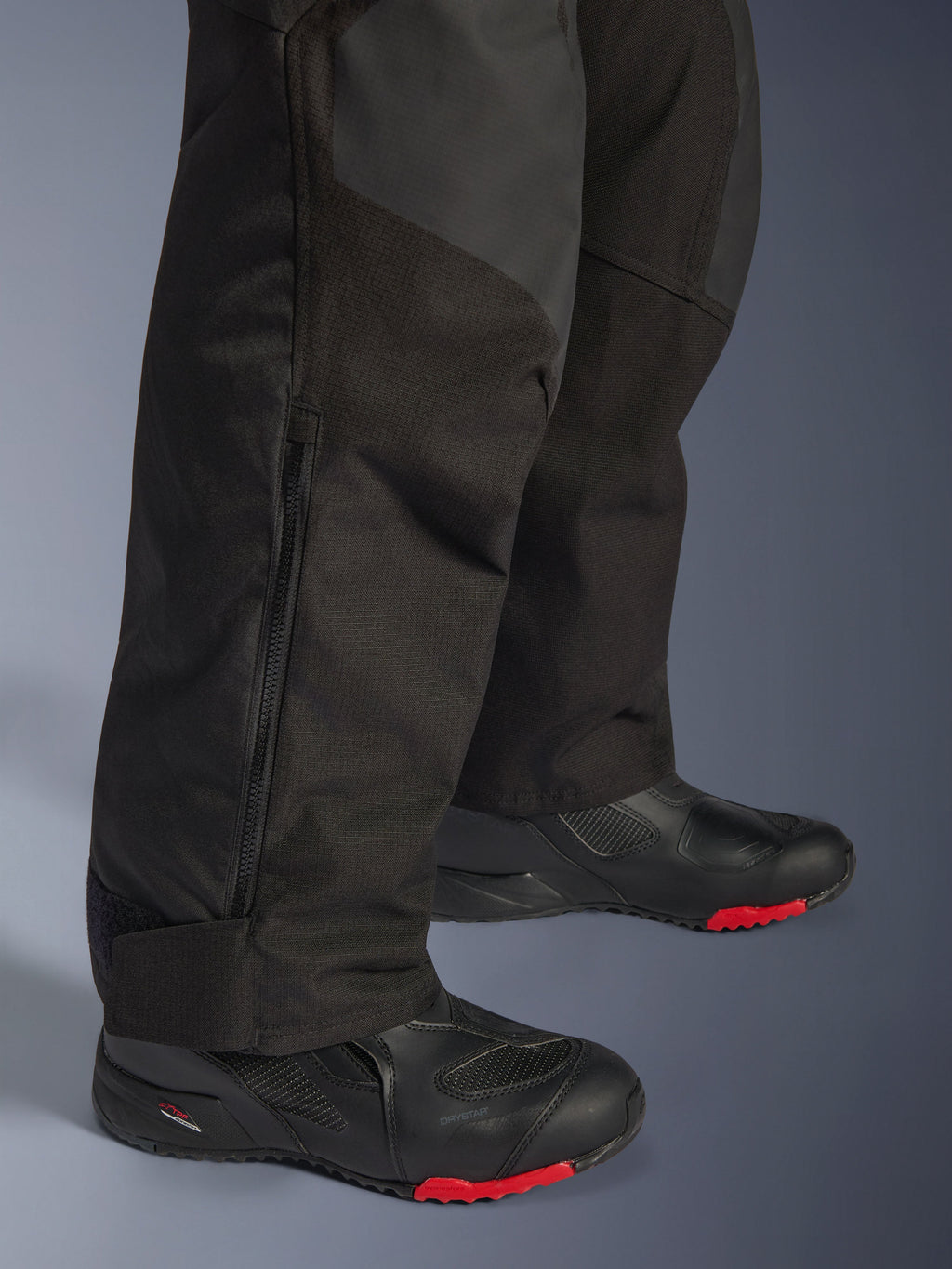 Alpinestars Andes V4 Drystar® Pants, pantaloni da moto adventure neri, primo piano della parte inferiore della gamba con dettaglio della cerniera laterale, tessuto resistente con rinforzi in ripstop, indossati con stivali neri con suole rosse
