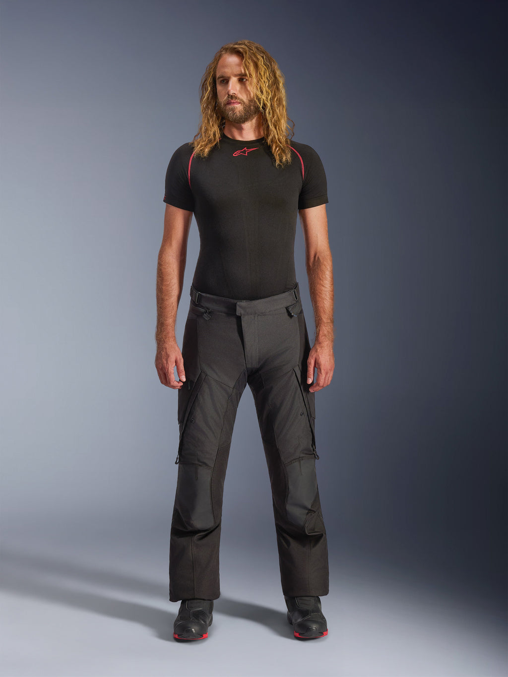 Modello che indossa i pantaloni Alpinestars Andes V4 Drystar® - Long in nero, pantaloni da moto adventure-touring con pannelli rinforzati sulle ginocchia e tasche cargo, abbinati a un base layer nero a maniche corte e stivali da moto neri