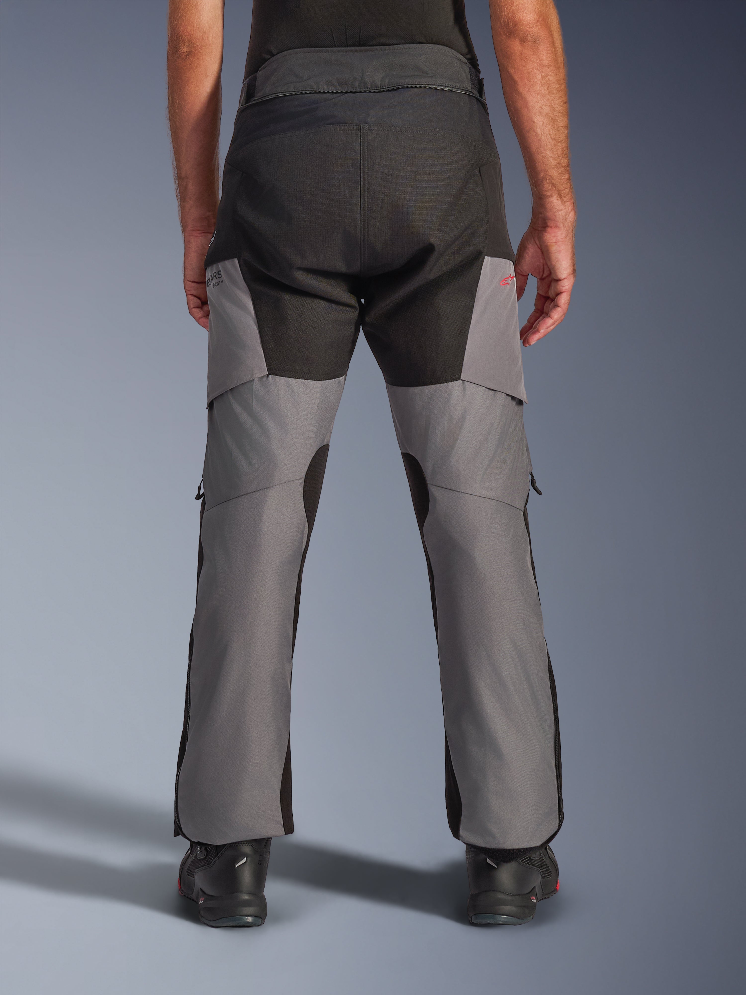Alpinestars Andes V4 Drystar® Pants, pantaloni da moto adventure, nero e grigio scuro, vista posteriore con seduta nera rinforzata e gambe grigie, piccolo logo rosso, abbinati a stivali da moto neri, struttura in tessuto resistente per touring in ogni condizione atmosferica.