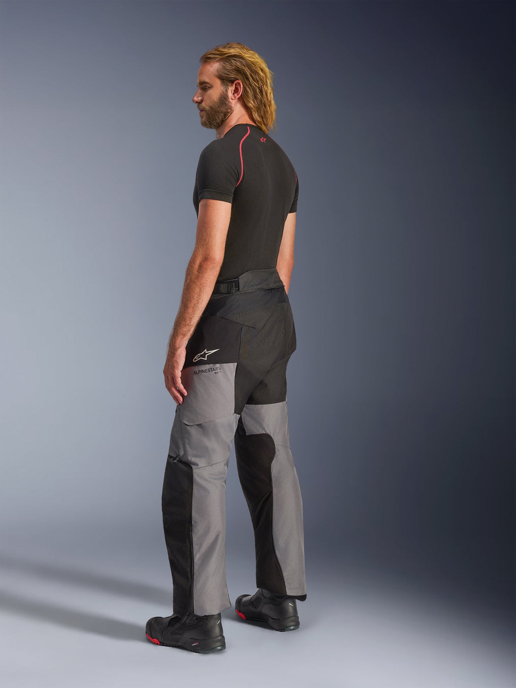 Alpinestars Andes V4 Drystar® Pants, pantaloni da moto adventure, nero e grigio scuro, indossati con base layer nero a maniche corte e stivali da moto neri, vista laterale e posteriore con design in tessuto multi-pannello e logo Alpinestars sulla coscia.