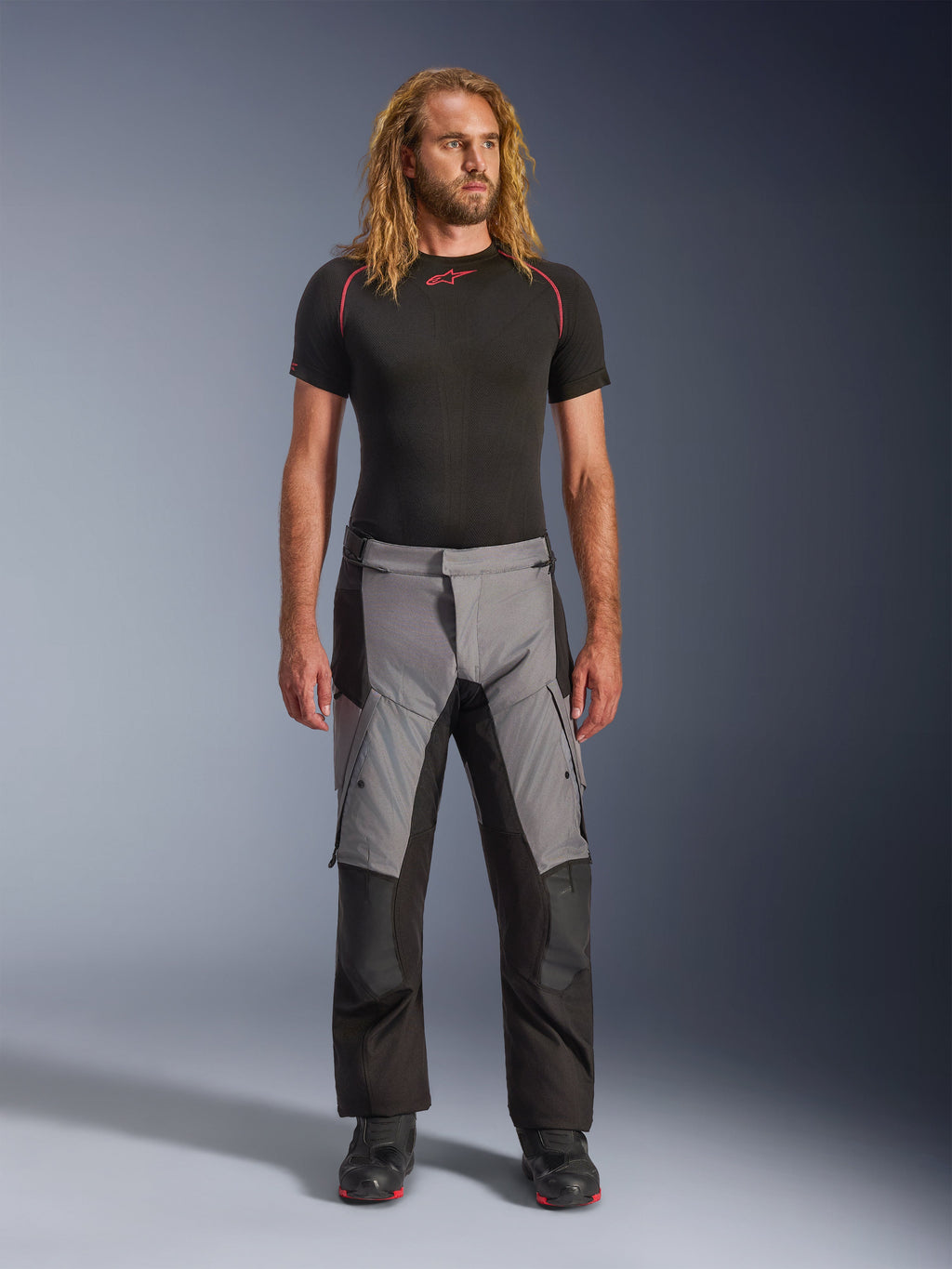 Pantaloni Alpinestars Andes V4 Drystar®, Nero e Grigio Scuro, pantaloni da moto adventure indossati da un modello con strato di base nero e stivali neri, caratterizzati da un design in tessuto multi-pannello con sezioni anteriori grigie e rinforzi neri, progettati per il touring in ogni condizione climatica e la guida su strada.