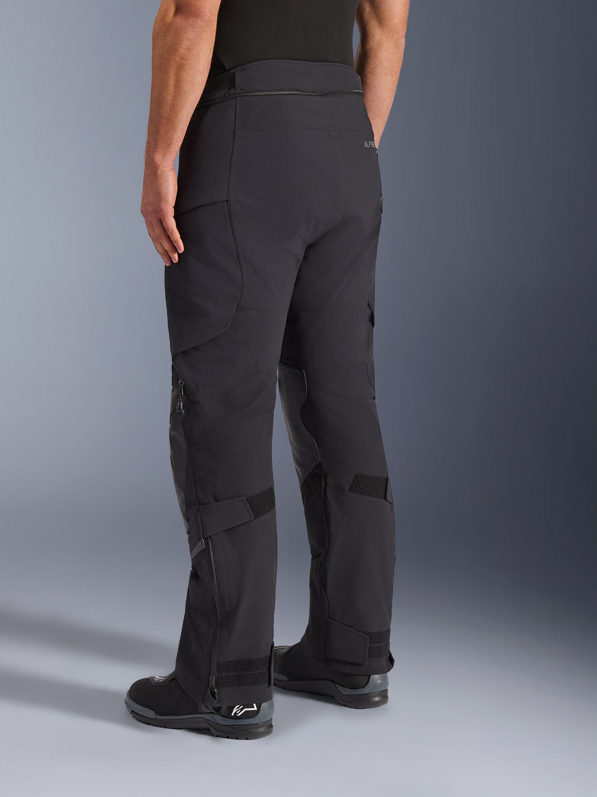 Pantaloni Alpinestars Andes Pro Drystar® XF, pantaloni adventure touring, nero e grigio scuro, vista posteriore a tre quarti che mostra la costruzione in tessuto, parte inferiore delle gambe con cerniera e cinturini di regolazione sui polpacci, e stivali da moto integrati