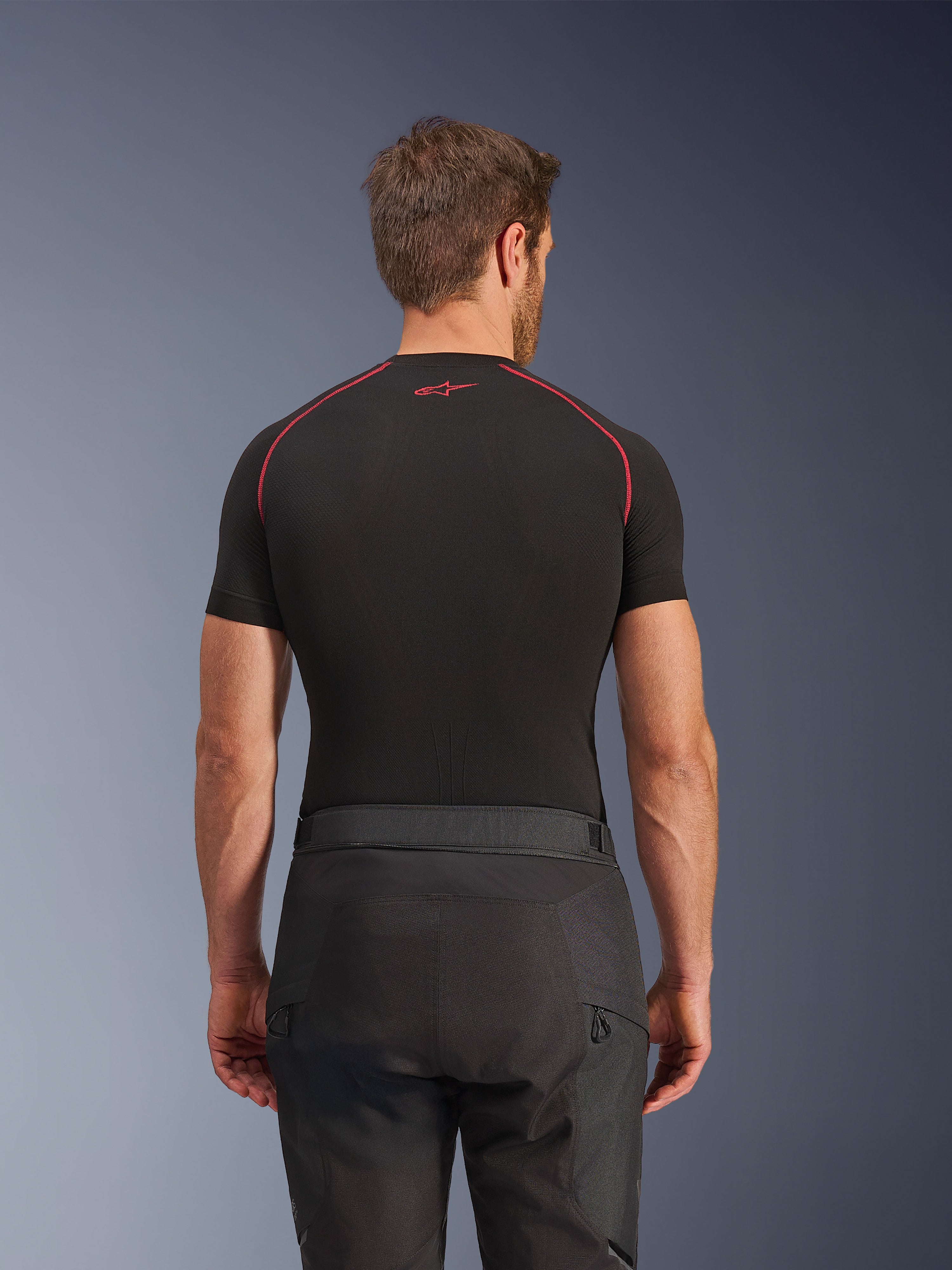 Pantaloni Alpinestars Cusco Drystar®, pantaloni da moto neri, vista posteriore di un pilota che indossa abbigliamento adventure touring con una maglia a compressione nera a maniche corte e cuciture rosse, robusta costruzione in tessuto ripstop e 600D.