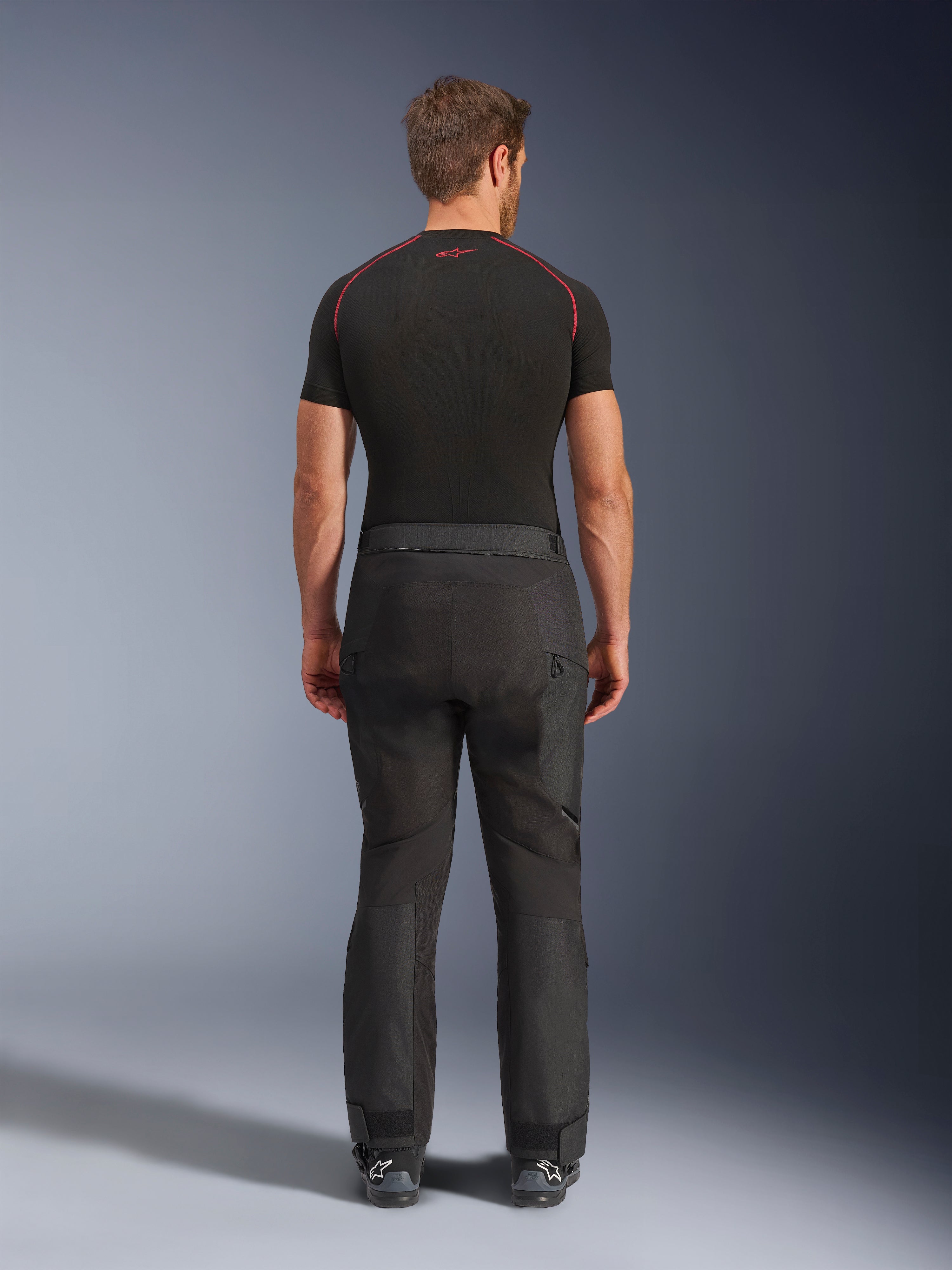 Pilota che indossa i pantaloni Alpinestars Cusco Drystar®, pantaloni da moto adventure neri, vista posteriore, abbinati a una maglia a compressione nera a maniche corte e stivali da moto neri, caratterizzati da un design in tessuto resistente con pannelli articolati per il touring