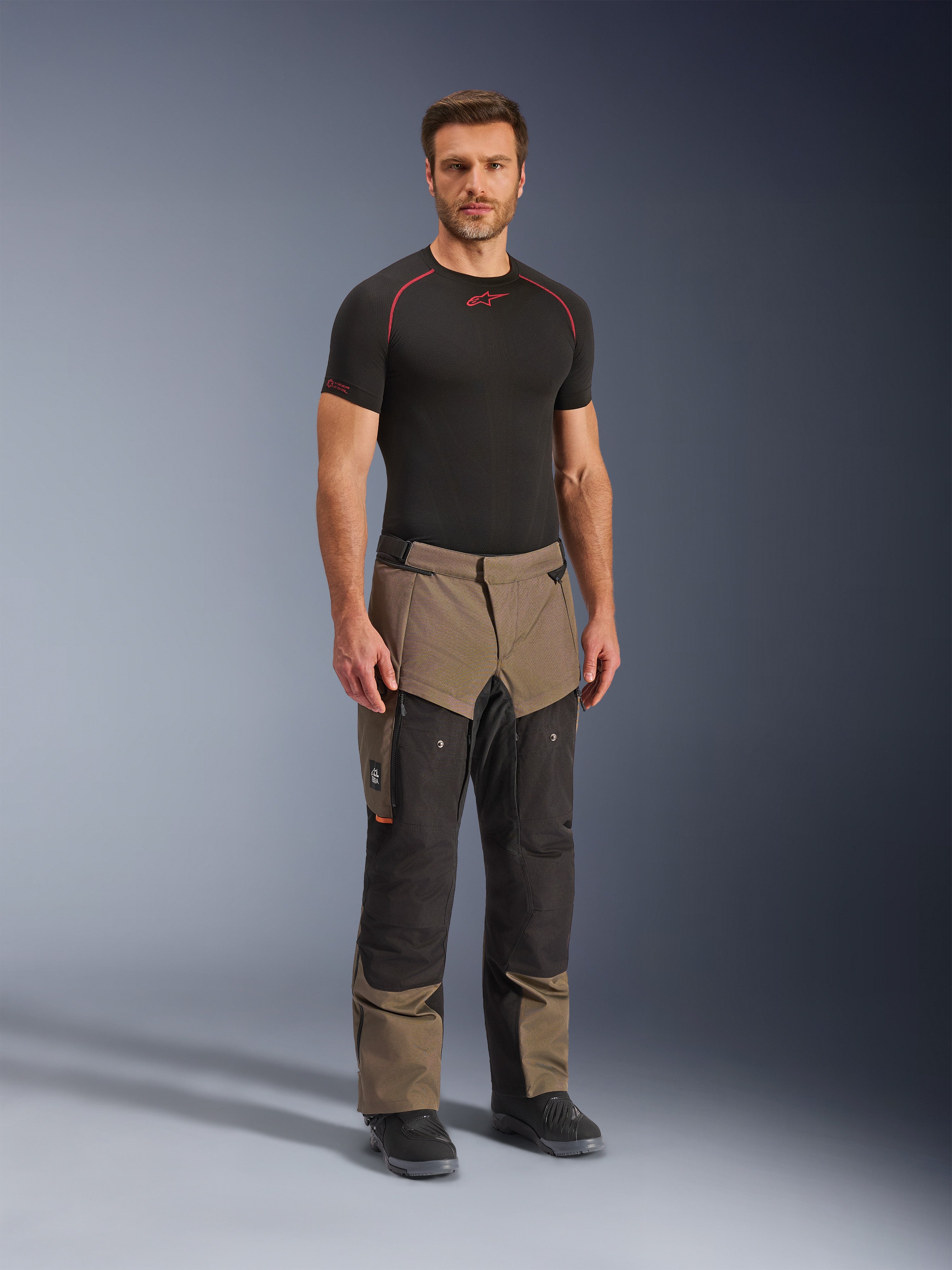 Pantaloni Alpinestars Cusco Drystar®, pantaloni da moto adventure, colore noce e nero, indossati da un modello con base layer nero e stivali, con ampie tasche cargo sulle cosce, zip di ventilazione e design bicolore robusto.