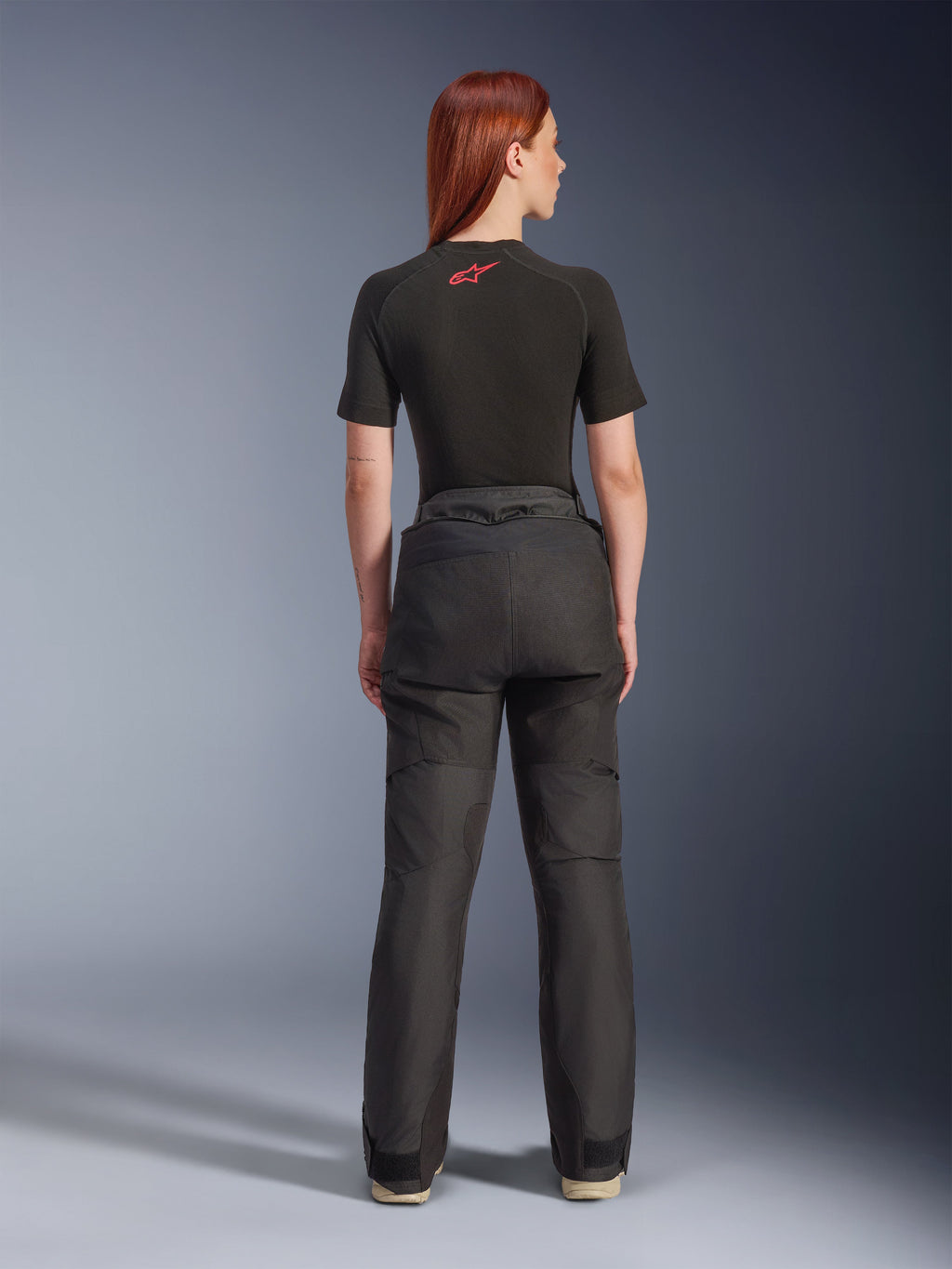 Vista posteriore di una modella che indossa i pantaloni Alpinestars Stella Andes V4 Drystar® neri, abbinati a un base layer nero a maniche corte con logo Alpinestars rosso, abbigliamento da moto adventure-touring con struttura in tessuto resistente.