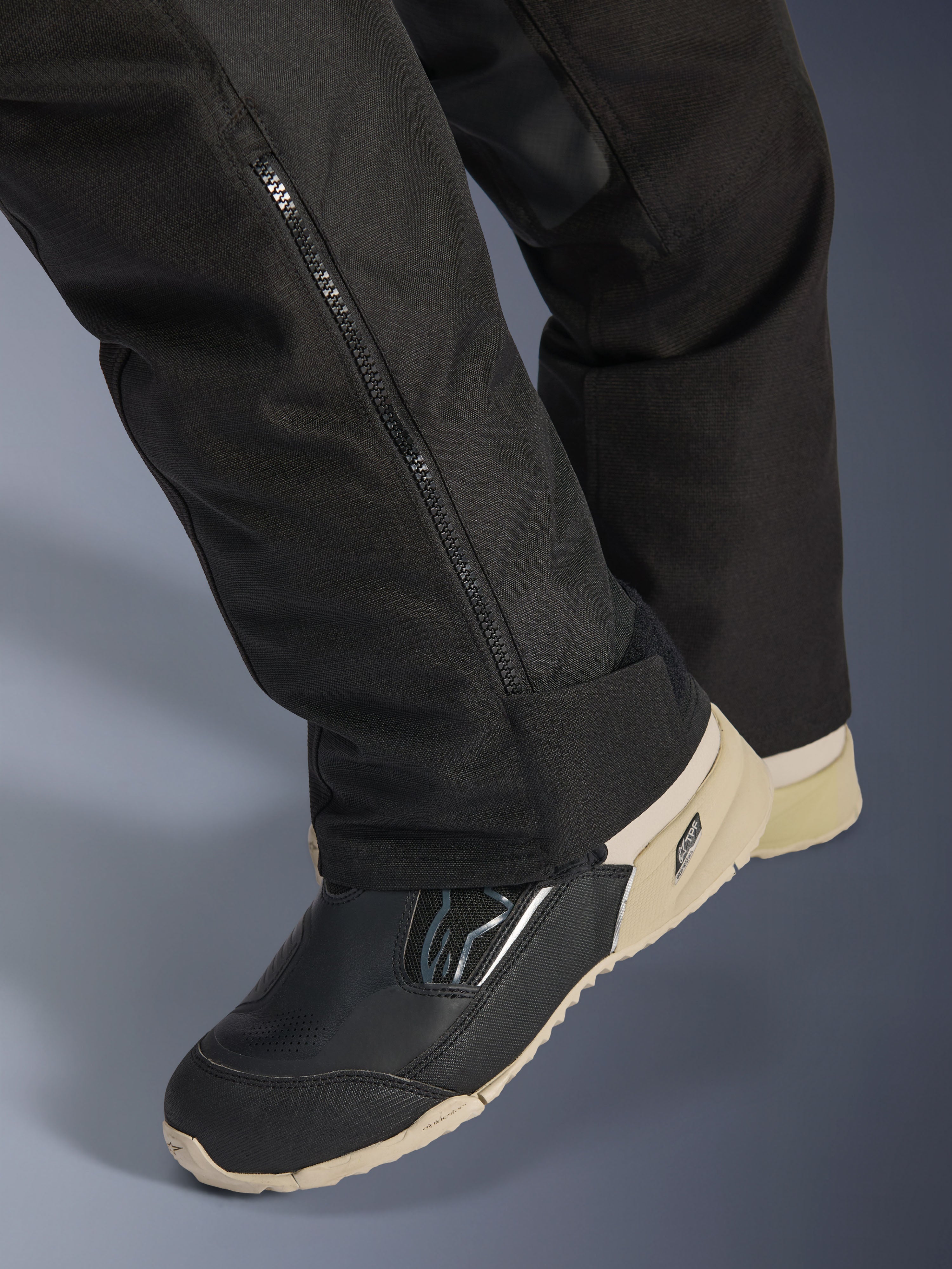 Alpinestars Stella Andes V4 Drystar® Pants, neri, primo piano della parte inferiore della gamba con cerniera laterale per la regolazione dello stivale, materiale tessile resistente, indossati con stivali da moto neri e sabbia, design funzionale per l'avventura