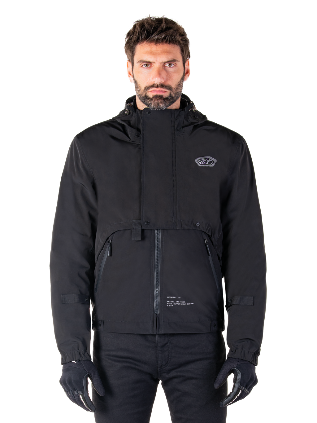 Alpinestars Mootant Waterproof Parka, giacca da moto nera con cappuccio dal design tecnico modulare, indossata da un modello con guanti neri, caratterizzata da un'esclusiva patta sul petto e tasche laterali con zip, stile urban e touring.