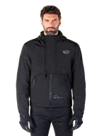 Alpinestars Mootant Waterproof Parka, giacca da moto nera con cappuccio dal design tecnico modulare, indossata da un modello con guanti neri, caratterizzata da un'esclusiva patta sul petto e tasche laterali con zip, stile urban e touring.
