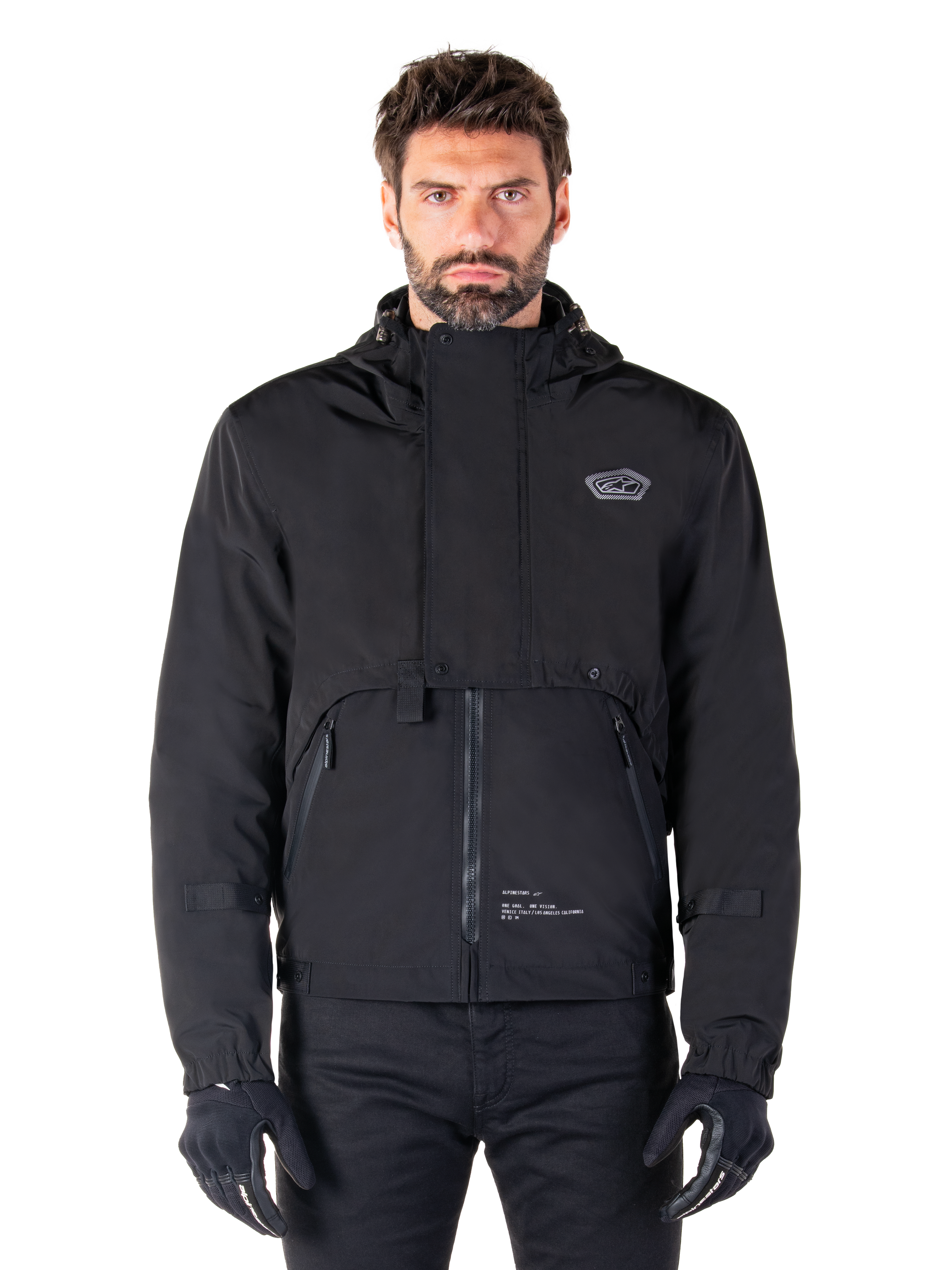 Alpinestars Mootant Waterproof Parka, giacca da moto nera con cappuccio dal design tecnico modulare, indossata da un modello con guanti neri, caratterizzata da un'esclusiva patta sul petto e tasche laterali con zip, stile urban e touring.