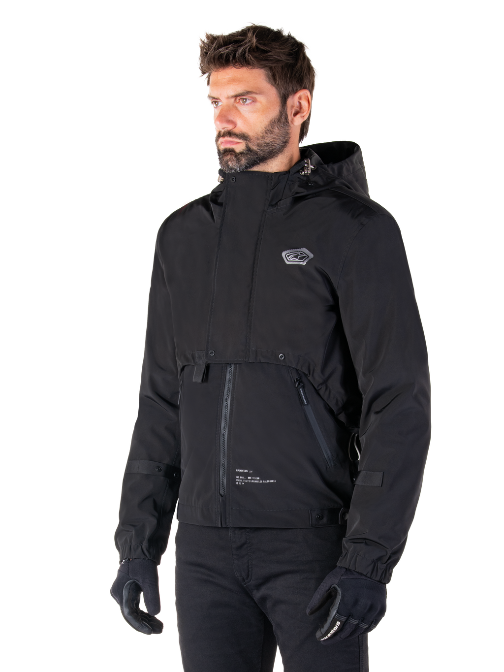 Alpinestars Parka Mootant Waterproof, giacca da moto modulare nera, indossata da un uomo con guanti e pantaloni neri, presenta cappuccio, cerniera frontale e design utilitaristico a strati, estetica urbana elegante con logo sul petto.