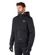 Alpinestars Parka Mootant Waterproof, giacca da moto modulare nera, indossata da un uomo con guanti e pantaloni neri, presenta cappuccio, cerniera frontale e design utilitaristico a strati, estetica urbana elegante con logo sul petto.