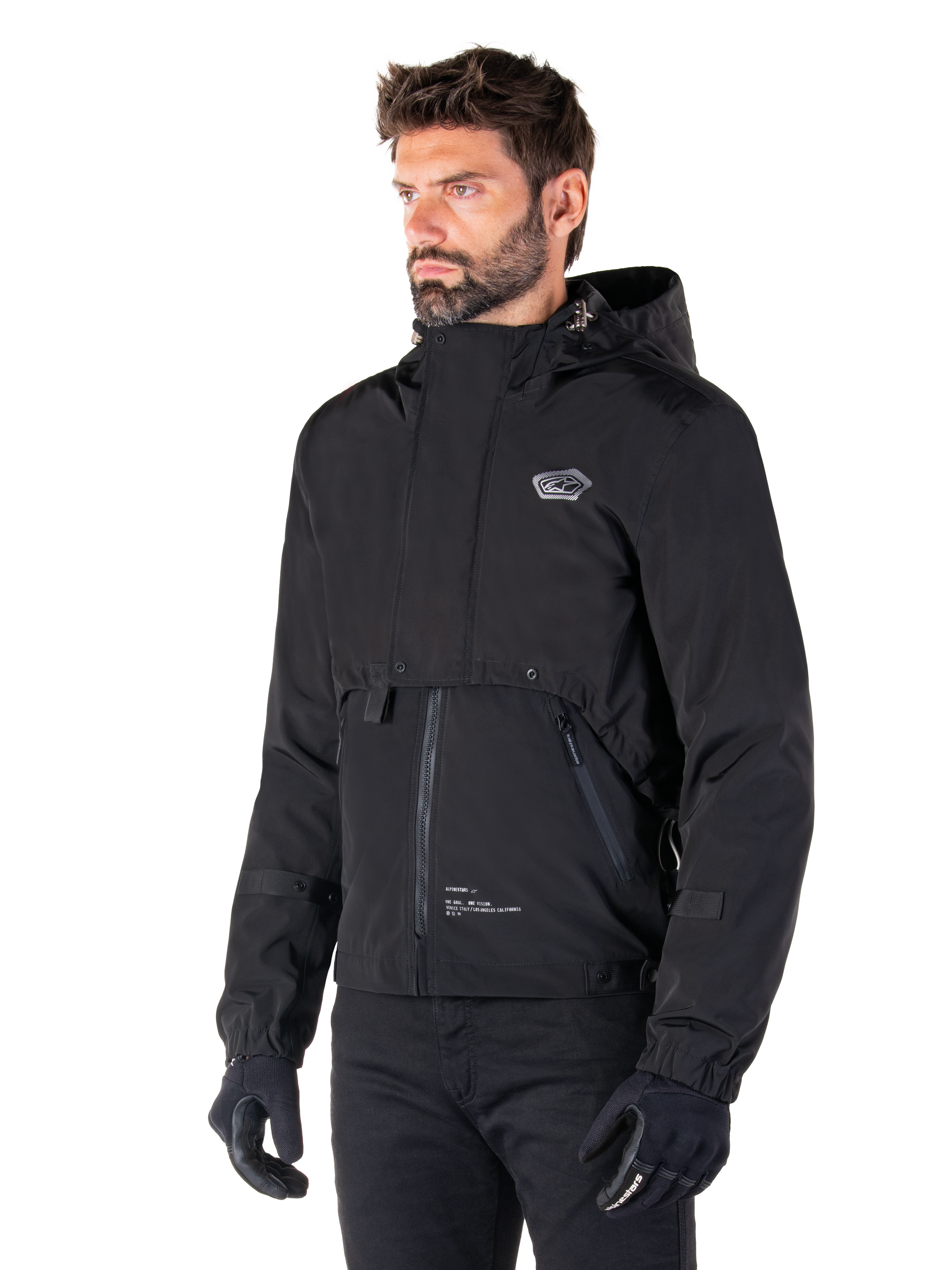 Alpinestars Parka Mootant Waterproof, giacca da moto modulare nera, indossata da un uomo con guanti e pantaloni neri, presenta cappuccio, cerniera frontale e design utilitaristico a strati, estetica urbana elegante con logo sul petto.