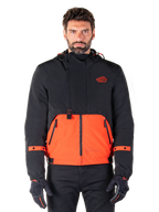Alpinestars Mootant Waterproof Parka, giacca da moto modulare in Spicy Orange e nero, indossata da un modello con guanti coordinati, design con cappuccio e pannelli bicolore, cerniera frontale e polsini regolabili, abbigliamento tecnico urbano contemporaneo