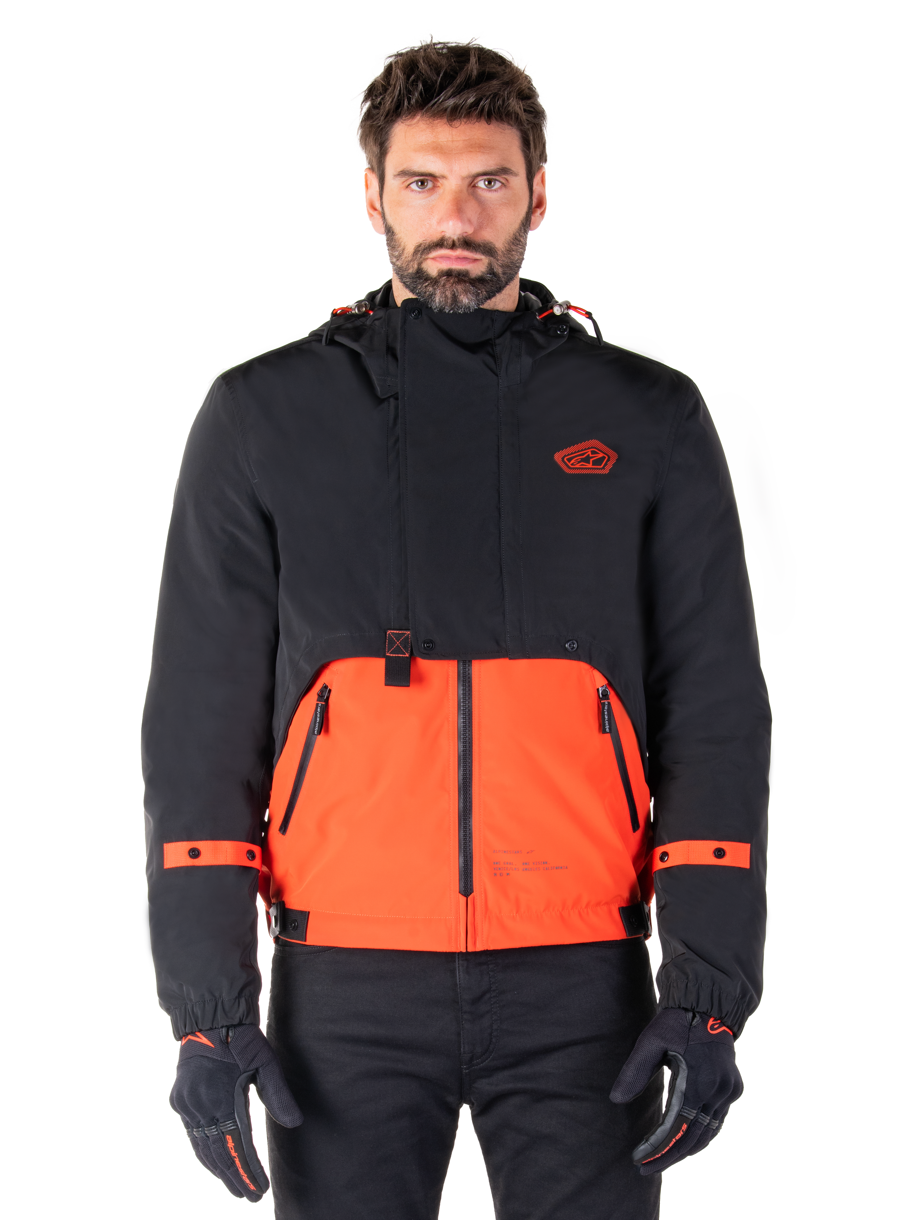 Alpinestars Mootant Waterproof Parka, giacca da moto modulare in Spicy Orange e nero, indossata da un modello con guanti coordinati, design con cappuccio e pannelli bicolore, cerniera frontale e polsini regolabili, abbigliamento tecnico urbano contemporaneo