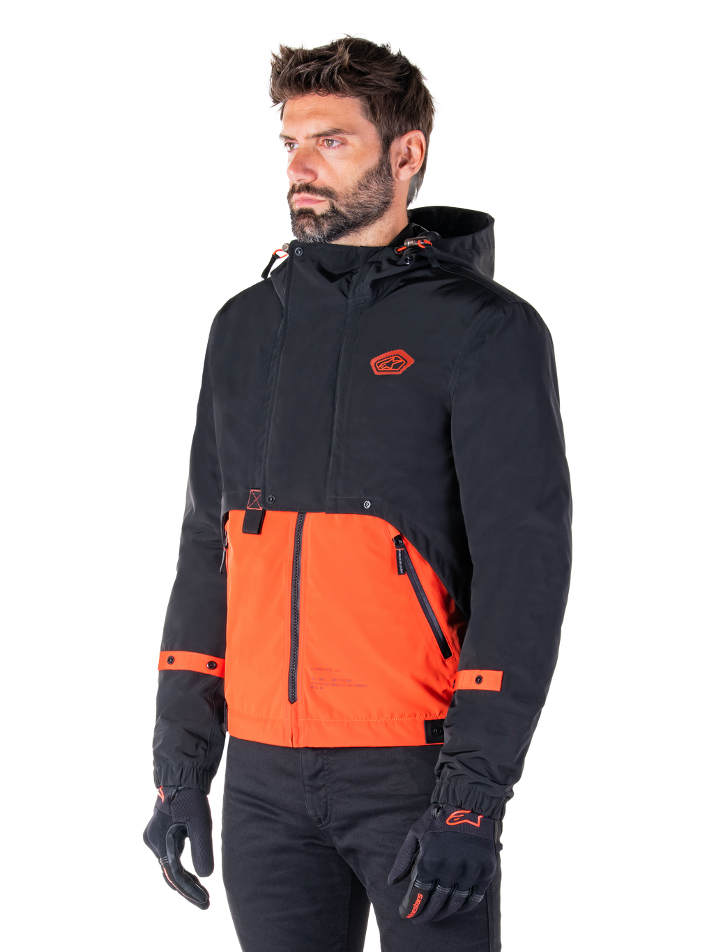 Uomo che indossa il parka impermeabile Alpinestars Mootant in Spicy Orange e nero, una giacca da moto modulare con strato superiore nero con cappuccio e sezione inferiore arancione brillante, abbinata a guanti da guida e pantaloni neri.